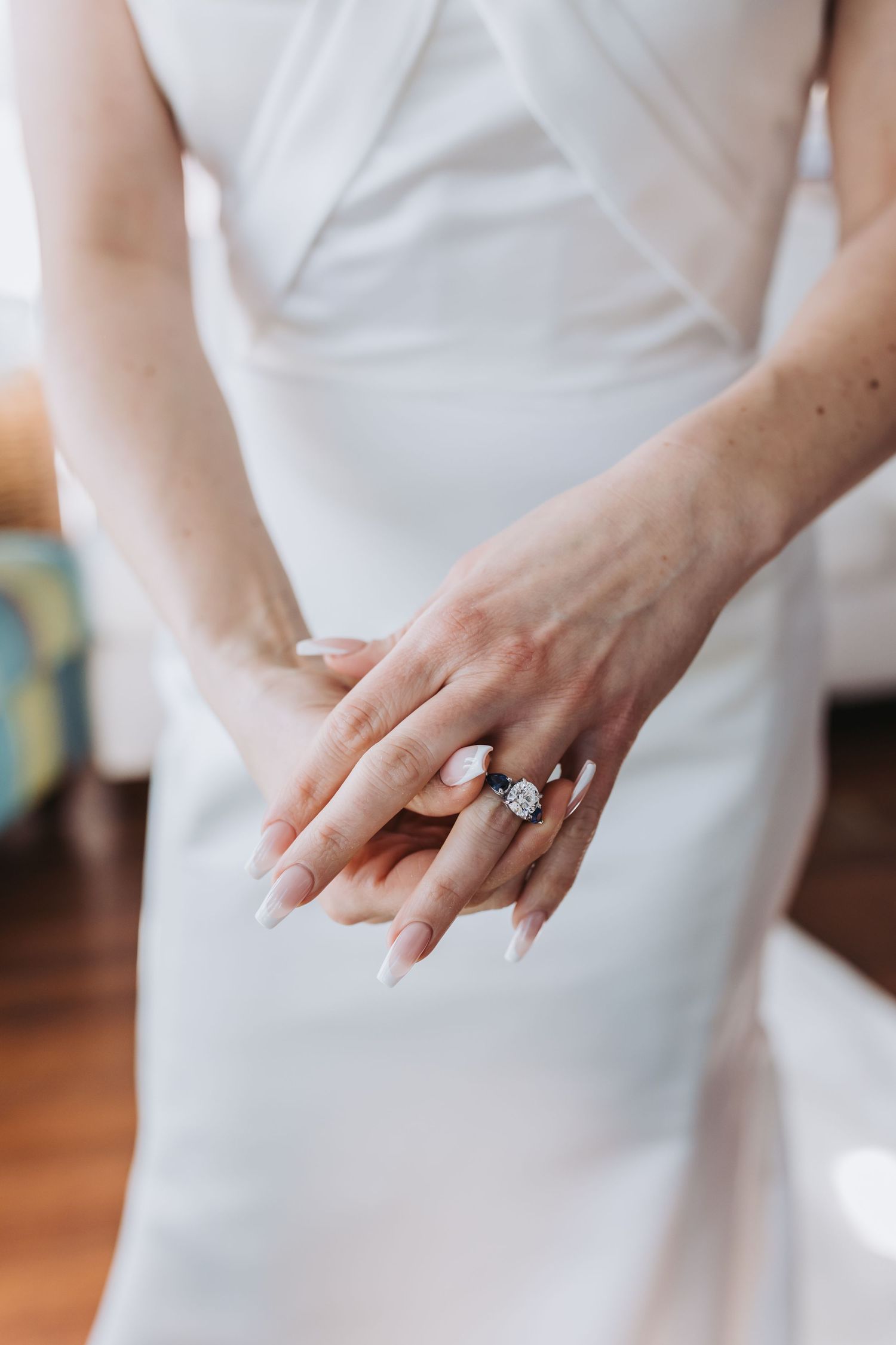 Close-up of bride\u2019s hands showcasing elegant diamond engagement ring against white wedding dress.