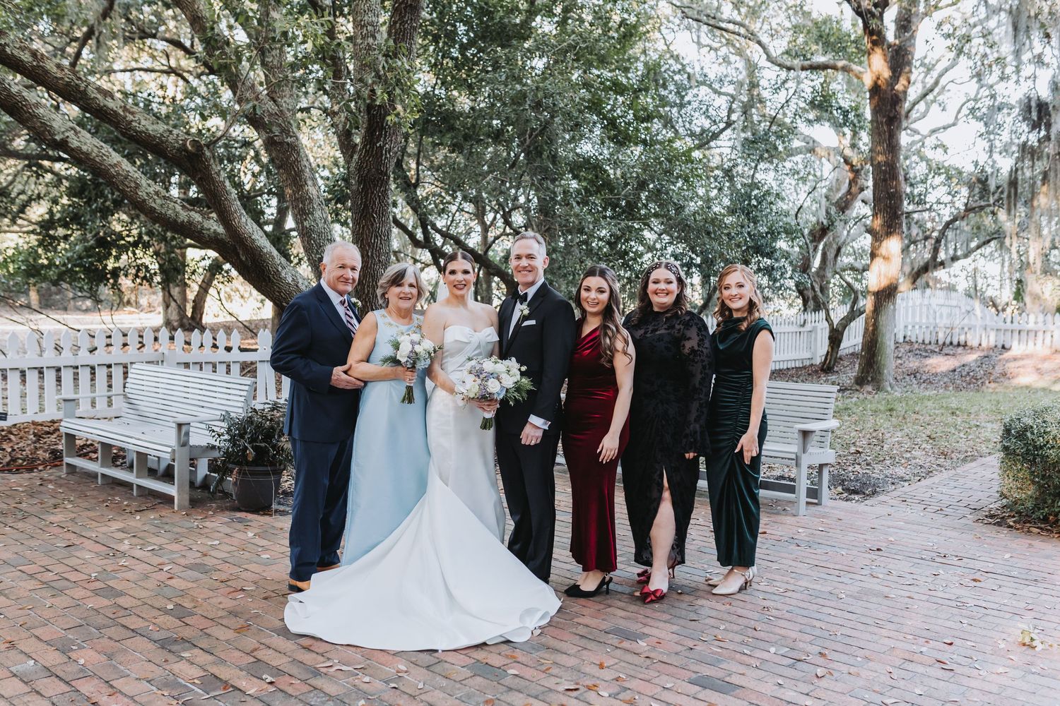 Wedding party of six poses together on brick pathway surrounded by oak trees and white fences.