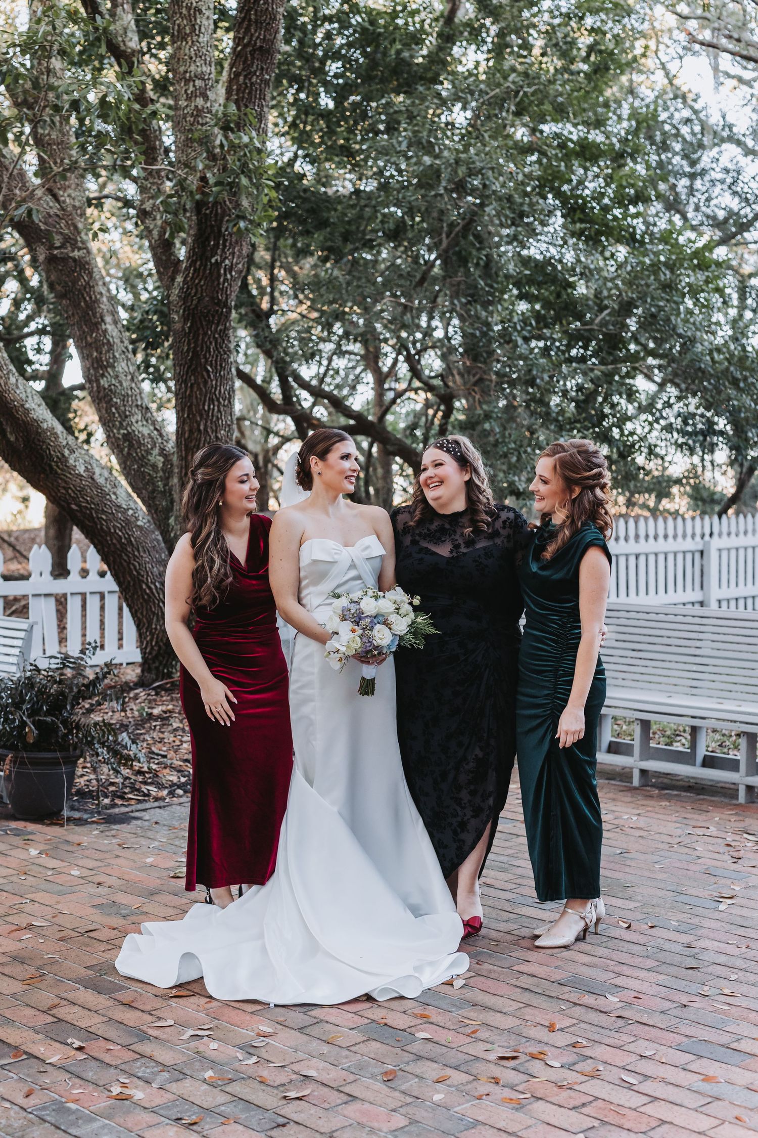 Bride in white gown poses with three bridesmaids in jewel-toned velvet dresses under oak trees on brick path.