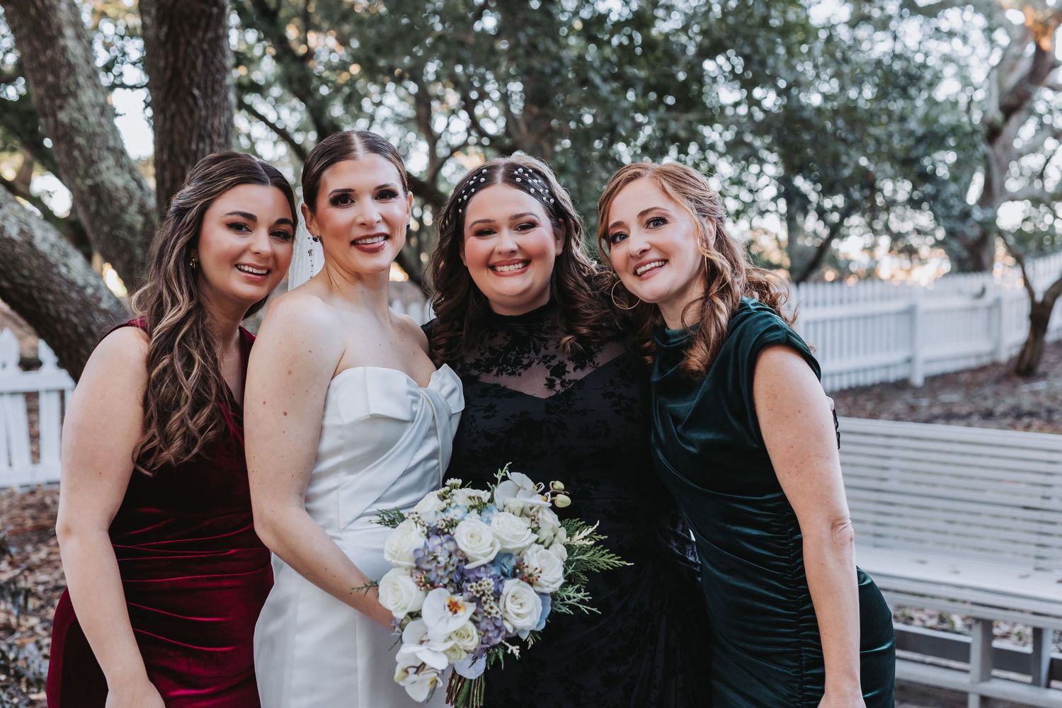 Bride with white bouquet poses joyfully with three bridesmaids in colorful dresses under oak trees.