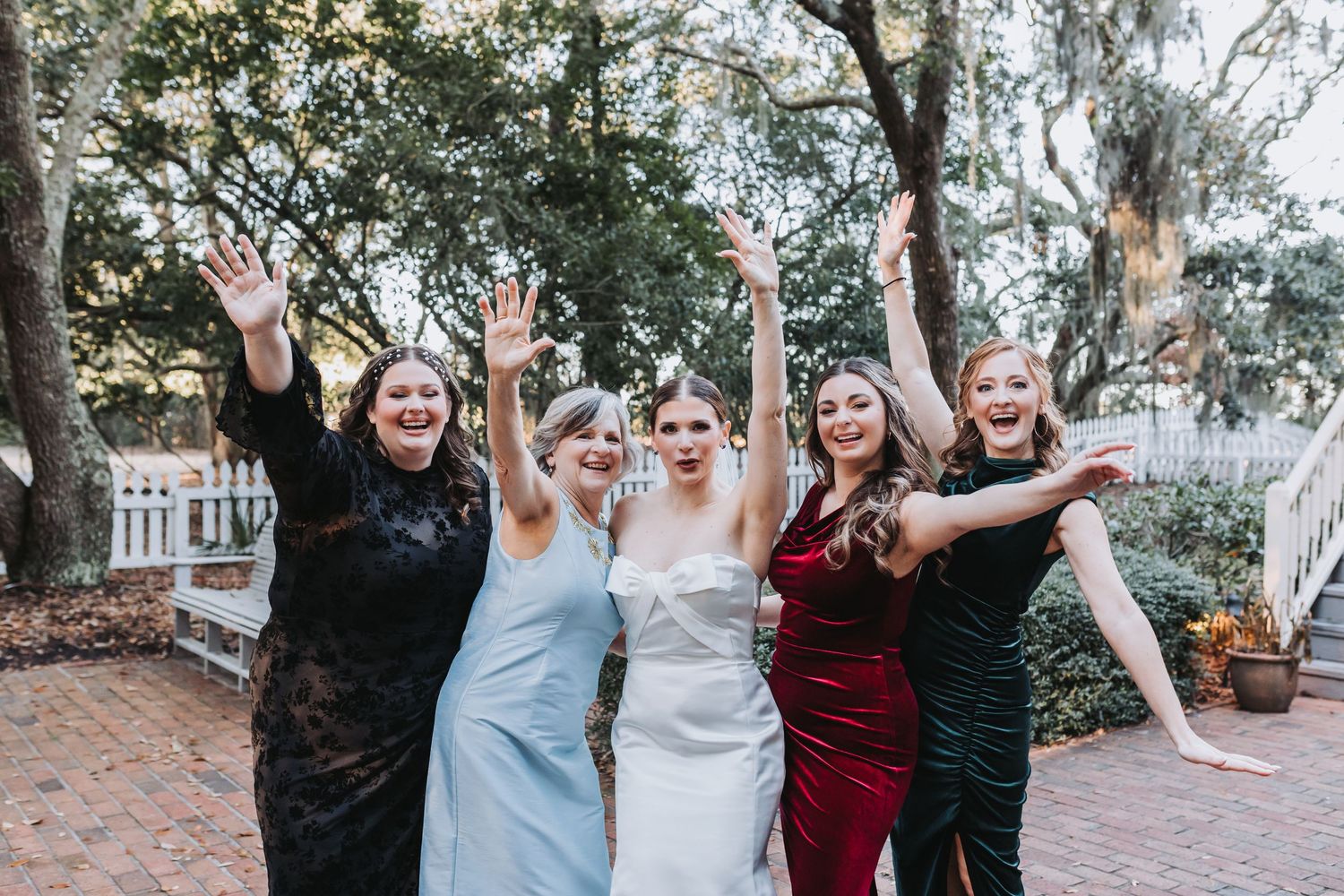Bride and four bridesmaids in colorful dresses celebrate with raised arms outdoors under large oak trees.