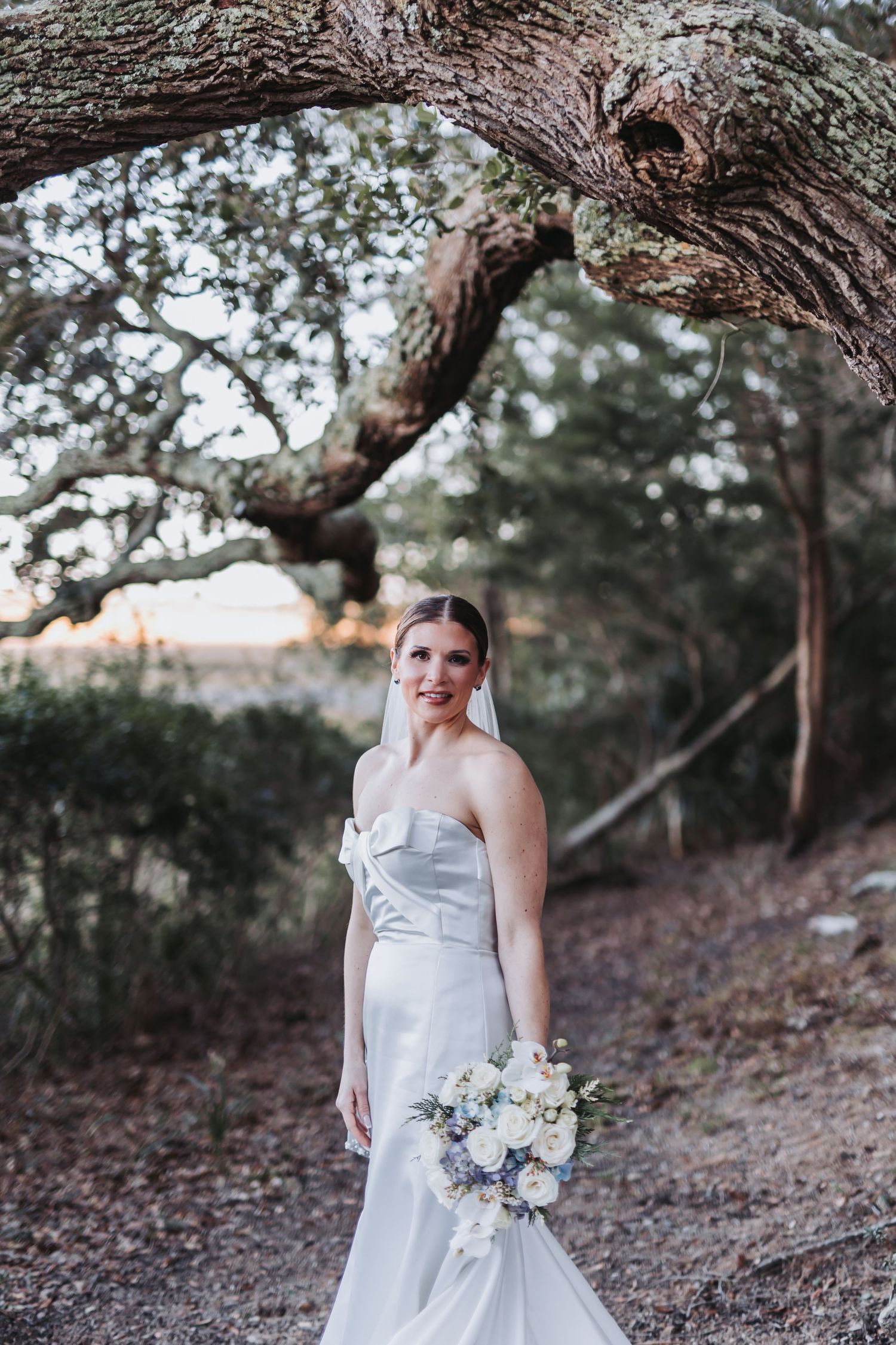 Bride in strapless white gown holding floral bouquet poses under a large oak tree in a wooded outdoor setting.