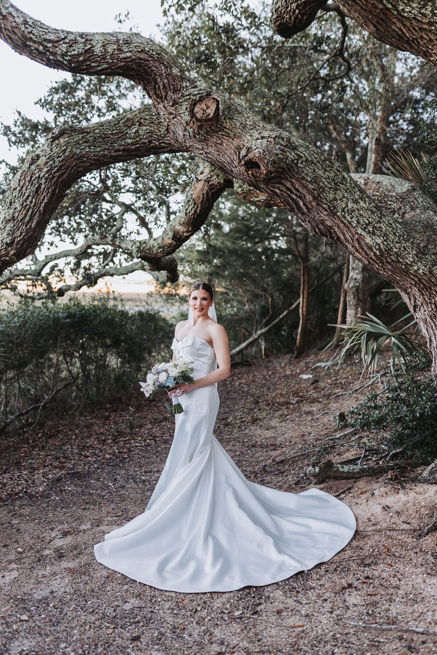 Elegant bride in strapless satin gown with long train holding bouquet beneath a dramatic oak tree.