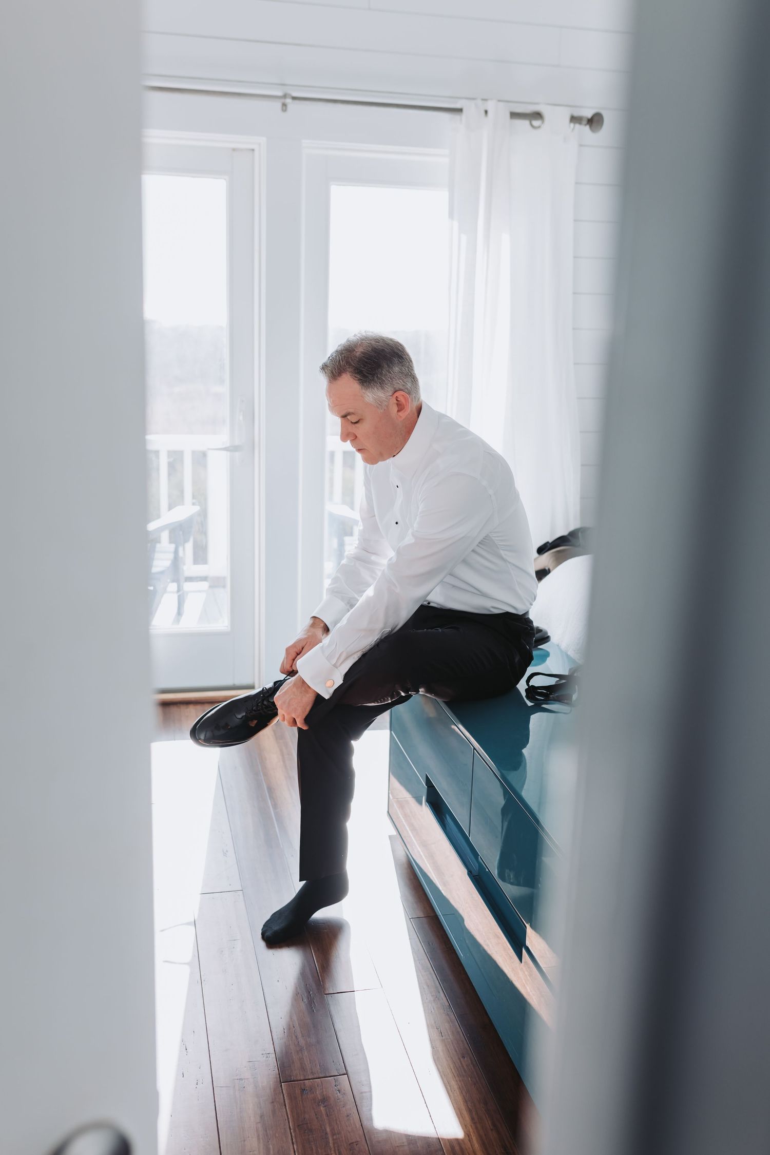 Mature groom in white dress shirt sitting on bed putting on black dress shoes before wedding ceremony.