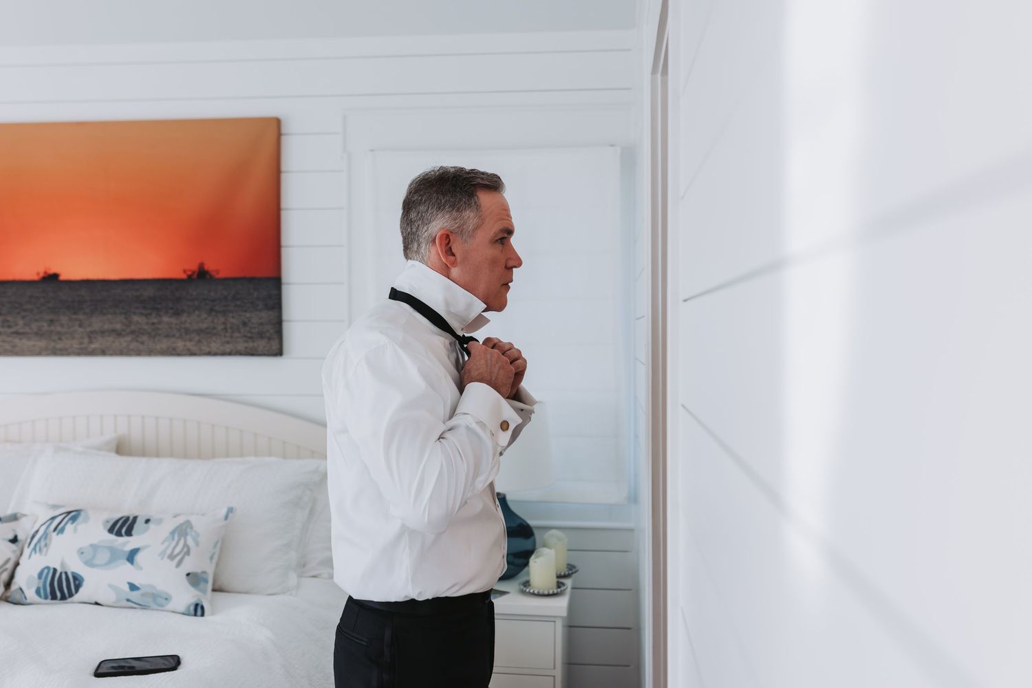 Groom in white dress shirt adjusting his bow tie in a bright bedroom before the wedding ceremony.
