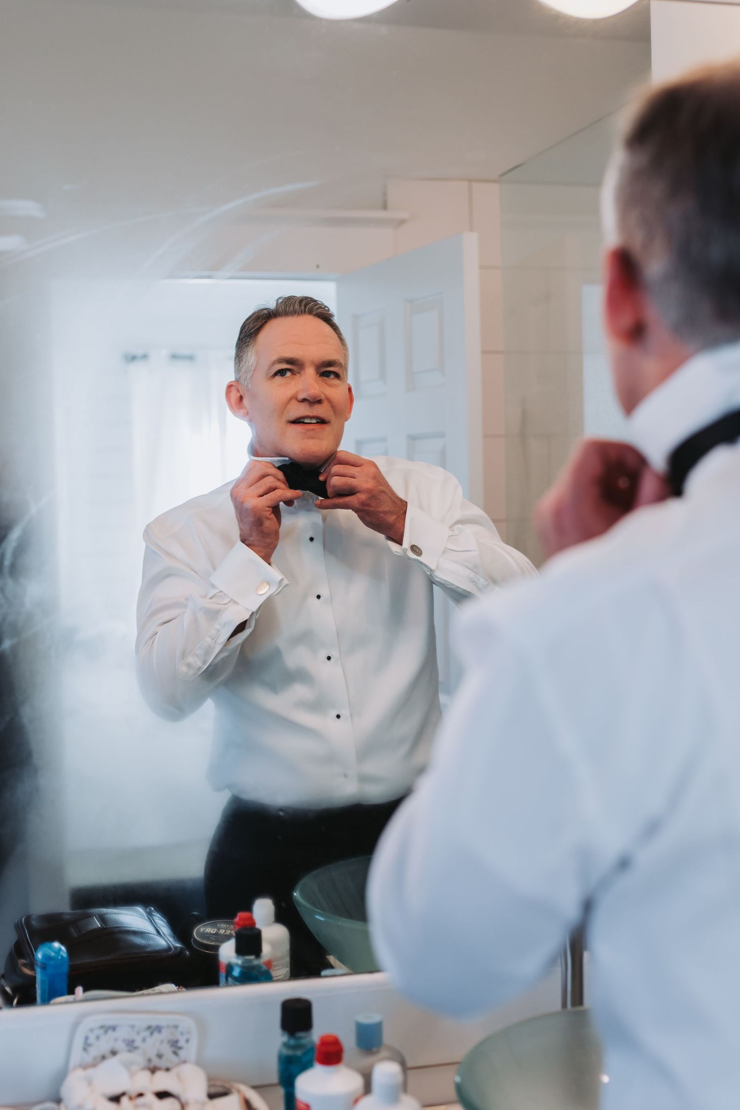Groom adjusts his bow tie while looking in the bathroom mirror while getting ready for wedding.
