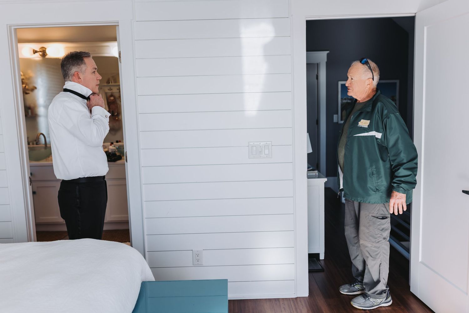 Groom in tuxedo adjusts his bow tie in a mirror while a man in a green jacket watches from the doorway.