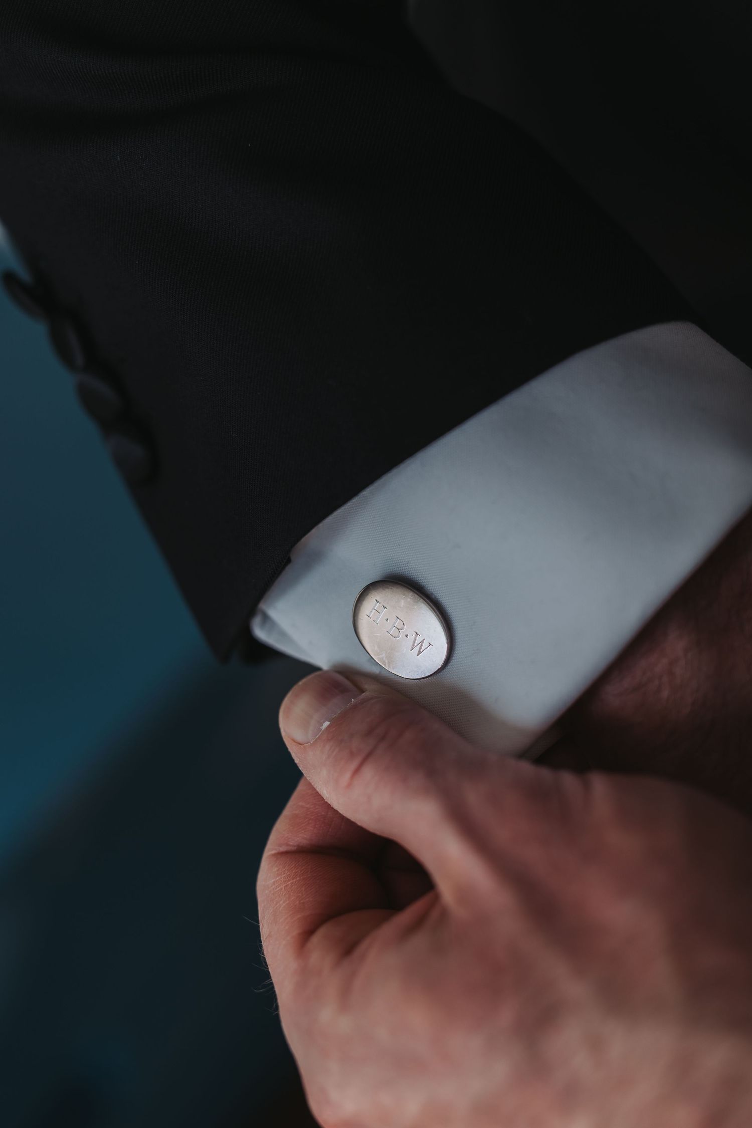 Close-up of groom fastening a silver cufflink on white dress shirt sleeve in black tuxedo jacket.