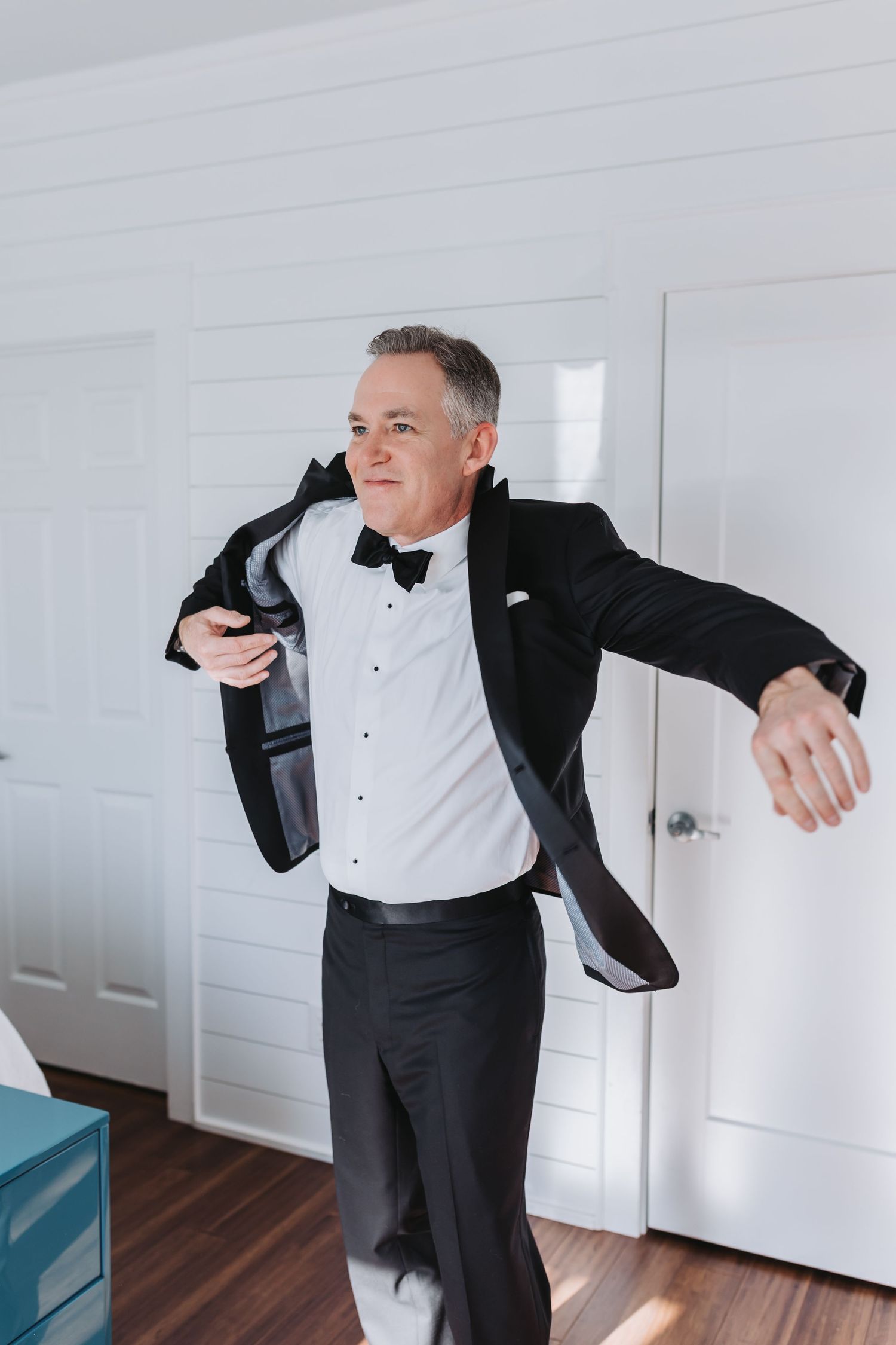 Dapper man in black tuxedo and bow tie putting on jacket in bright white room.
