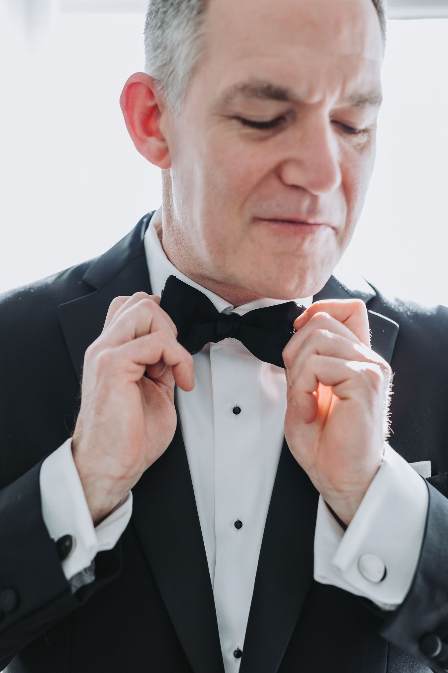 A groom in a black tuxedo adjusts his bow tie while looking down before his wedding ceremony.