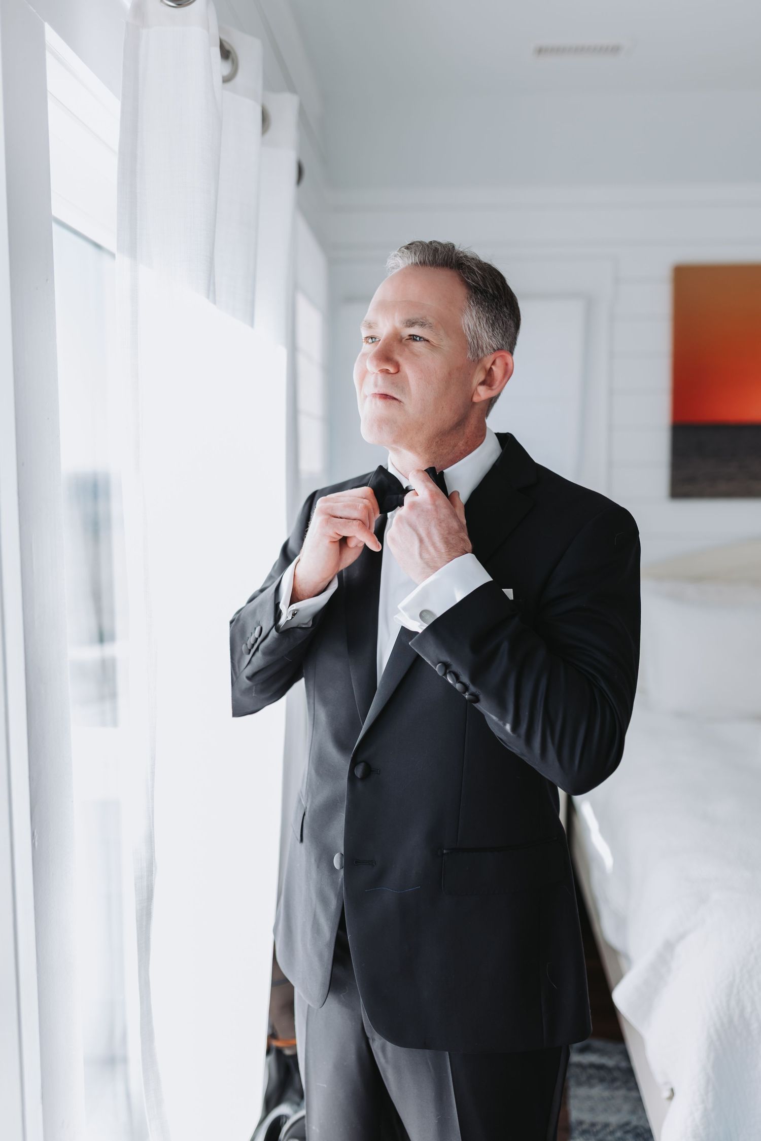 Distinguished groom in black tuxedo adjusting his tie while standing near a bright hotel window.