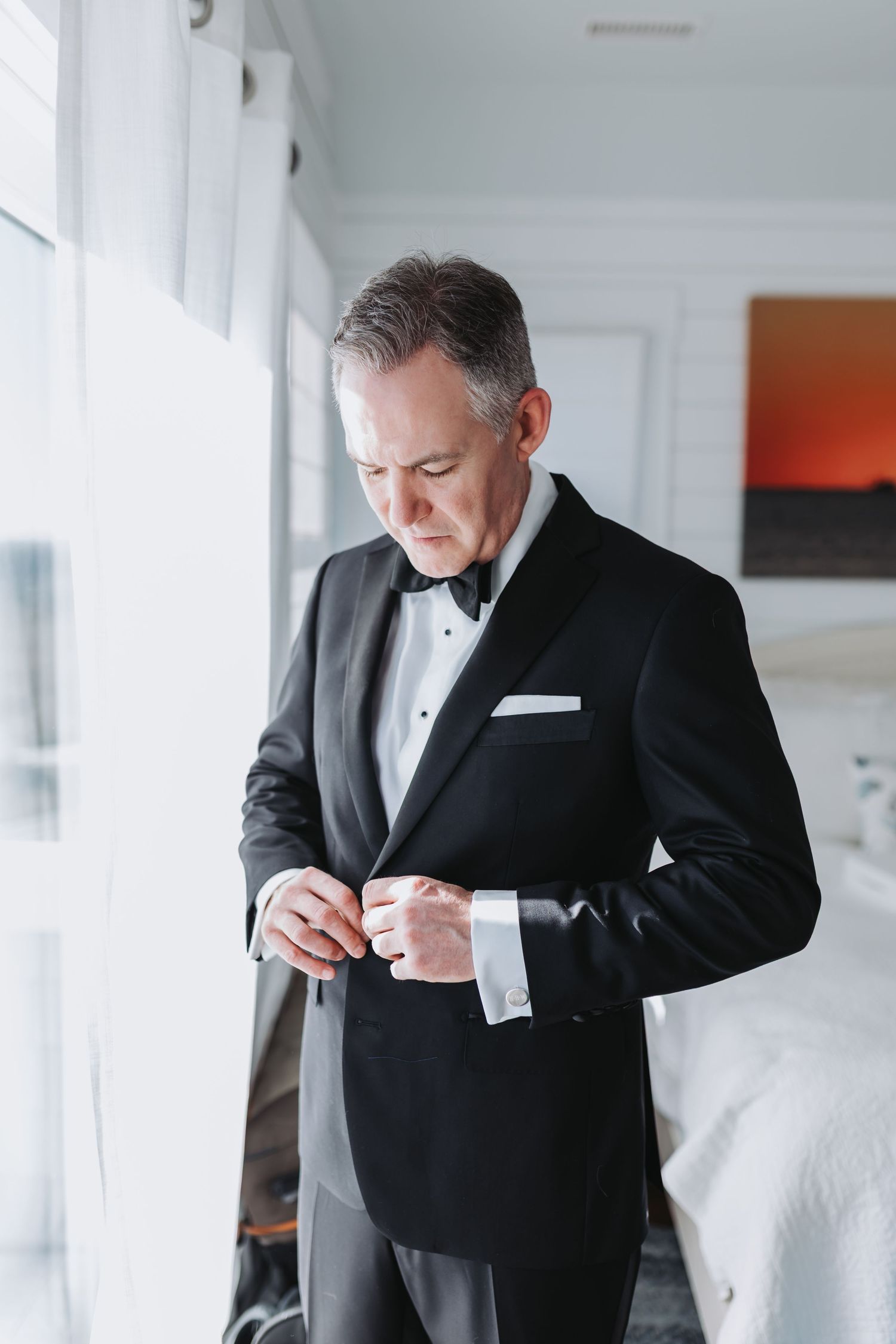 Dapper groom in black tuxedo fastening cufflinks while standing by a window on his wedding day.