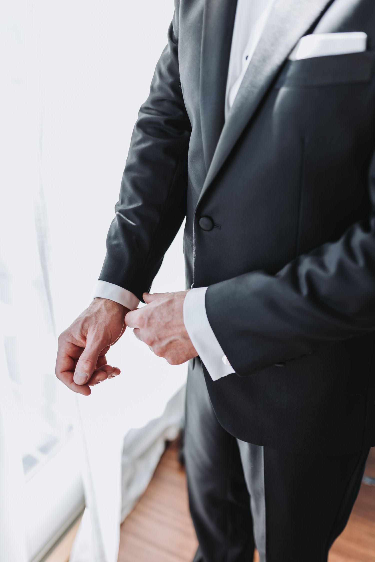Close-up of groom in black tuxedo adjusting his cufflinks near a bright window.