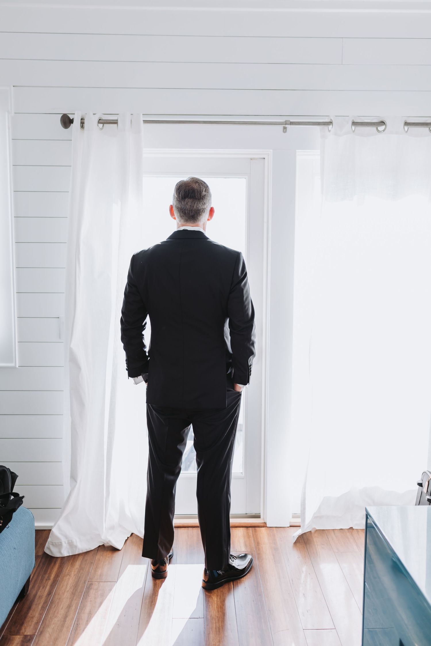Groom in black tuxedo stands looking out bright white curtained doorway on wedding day.