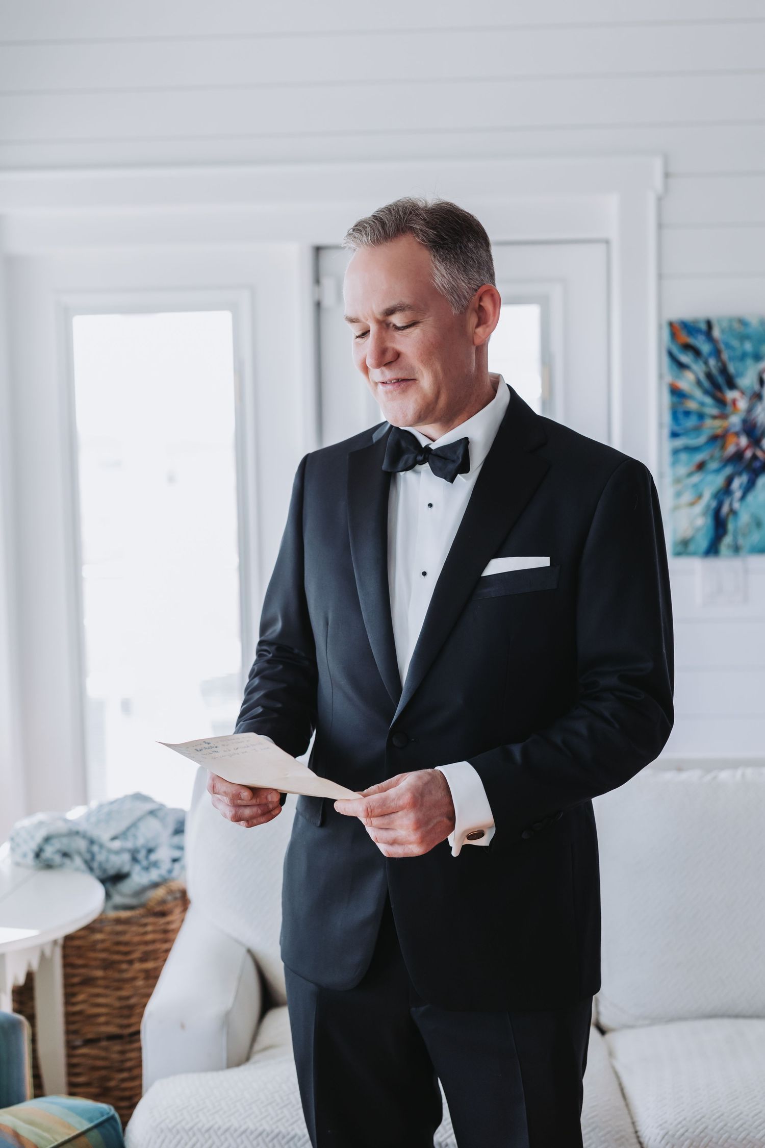 Dapper groom in black tuxedo and bow tie reads a heartfelt letter before his wedding ceremony.