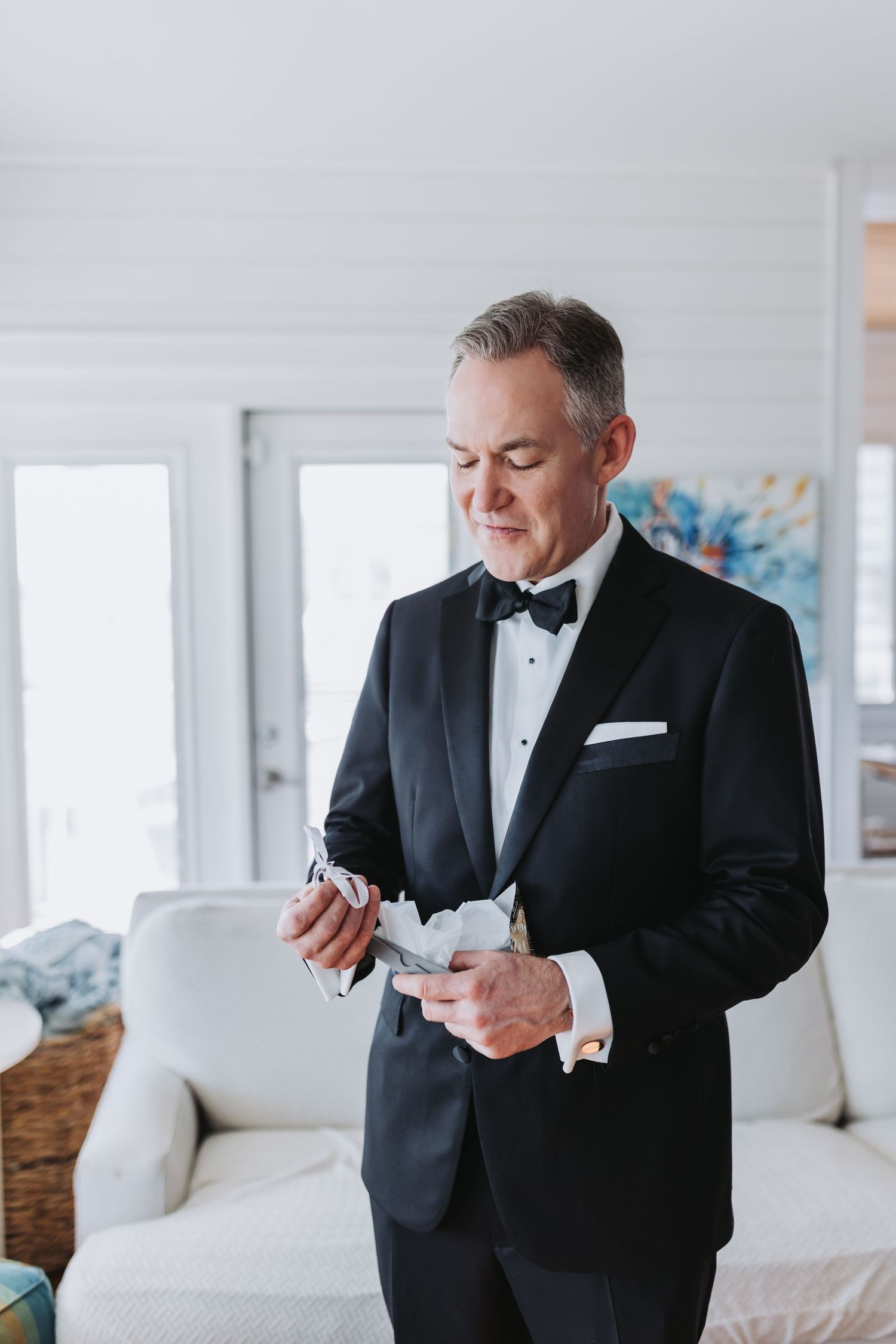 Groom in classic black tuxedo opening a gift box while standing in a bright white room.