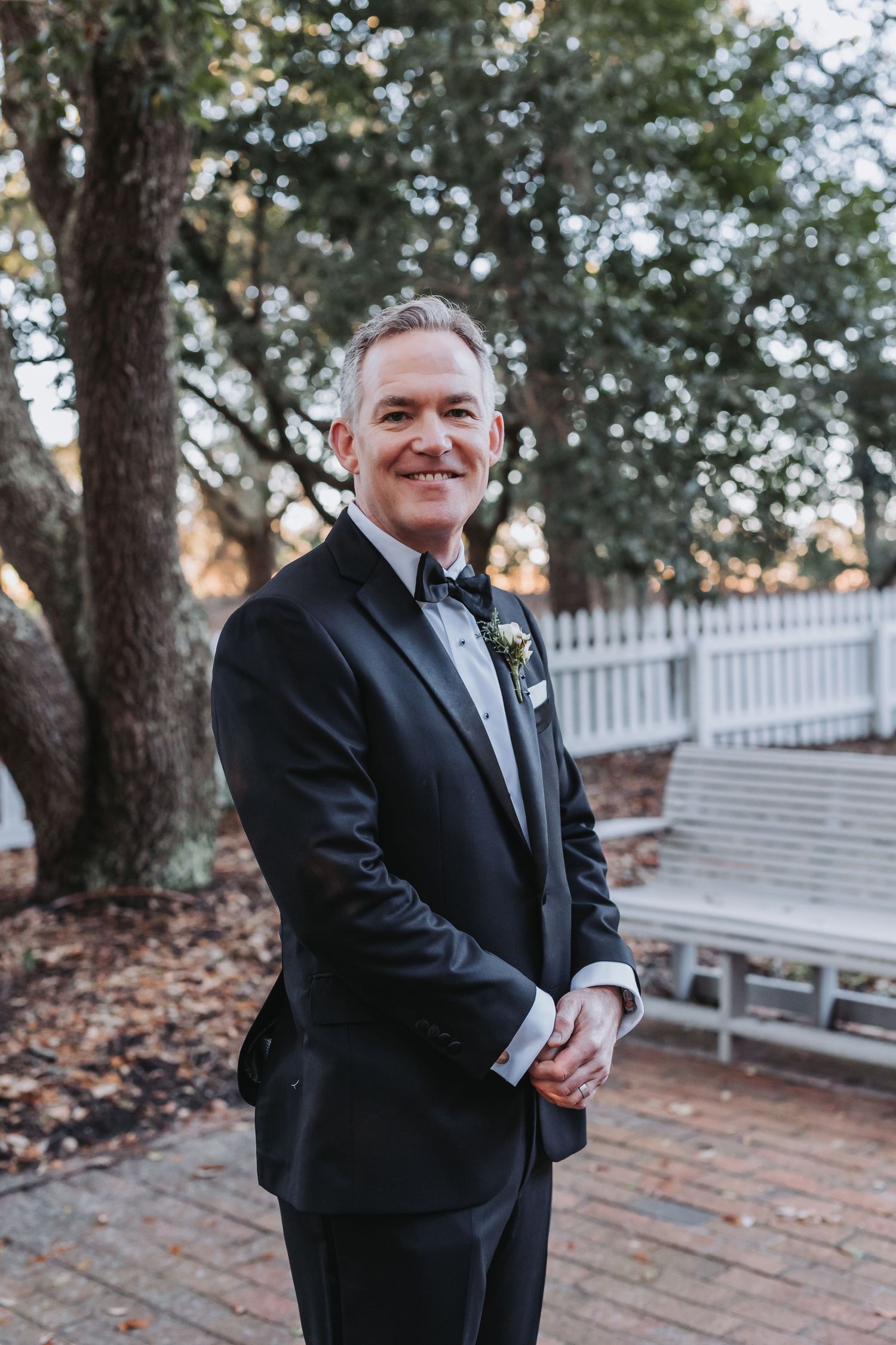 Groom in black tuxedo with bow tie and boutonniere smiling outdoors near white fence and oak trees.
