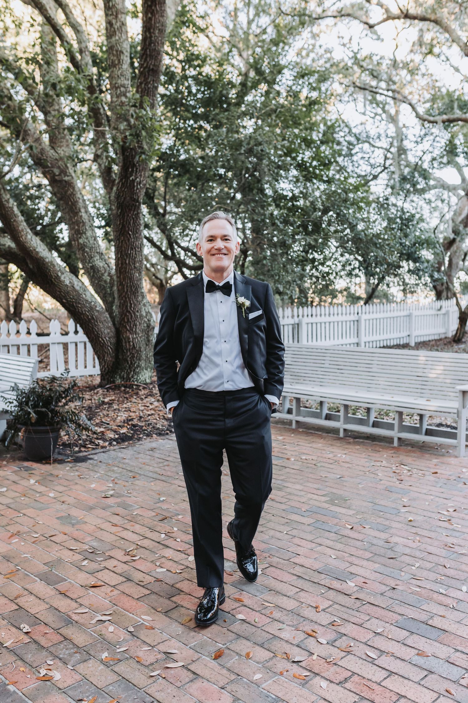 Smiling groom in classic black tuxedo walks confidently on brick path beneath large oak trees.