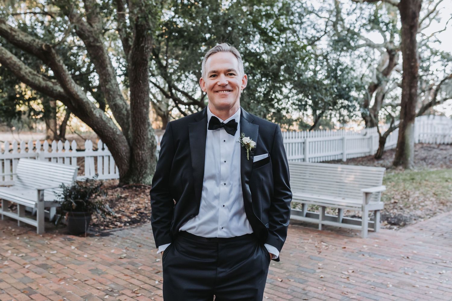 Smiling groom in a black tuxedo with boutonniere stands outdoors near benches and oak trees at a wedding venue.
