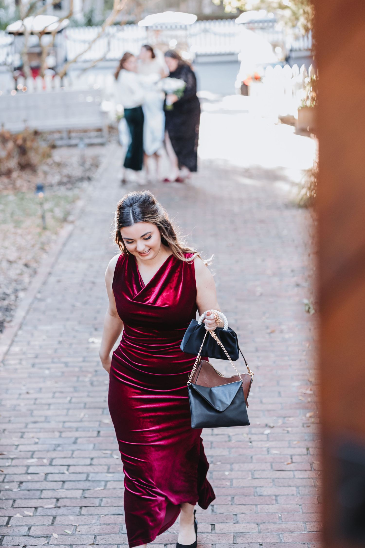 Woman in red velvet dress carrying black handbag walking on brick path at wedding venue.