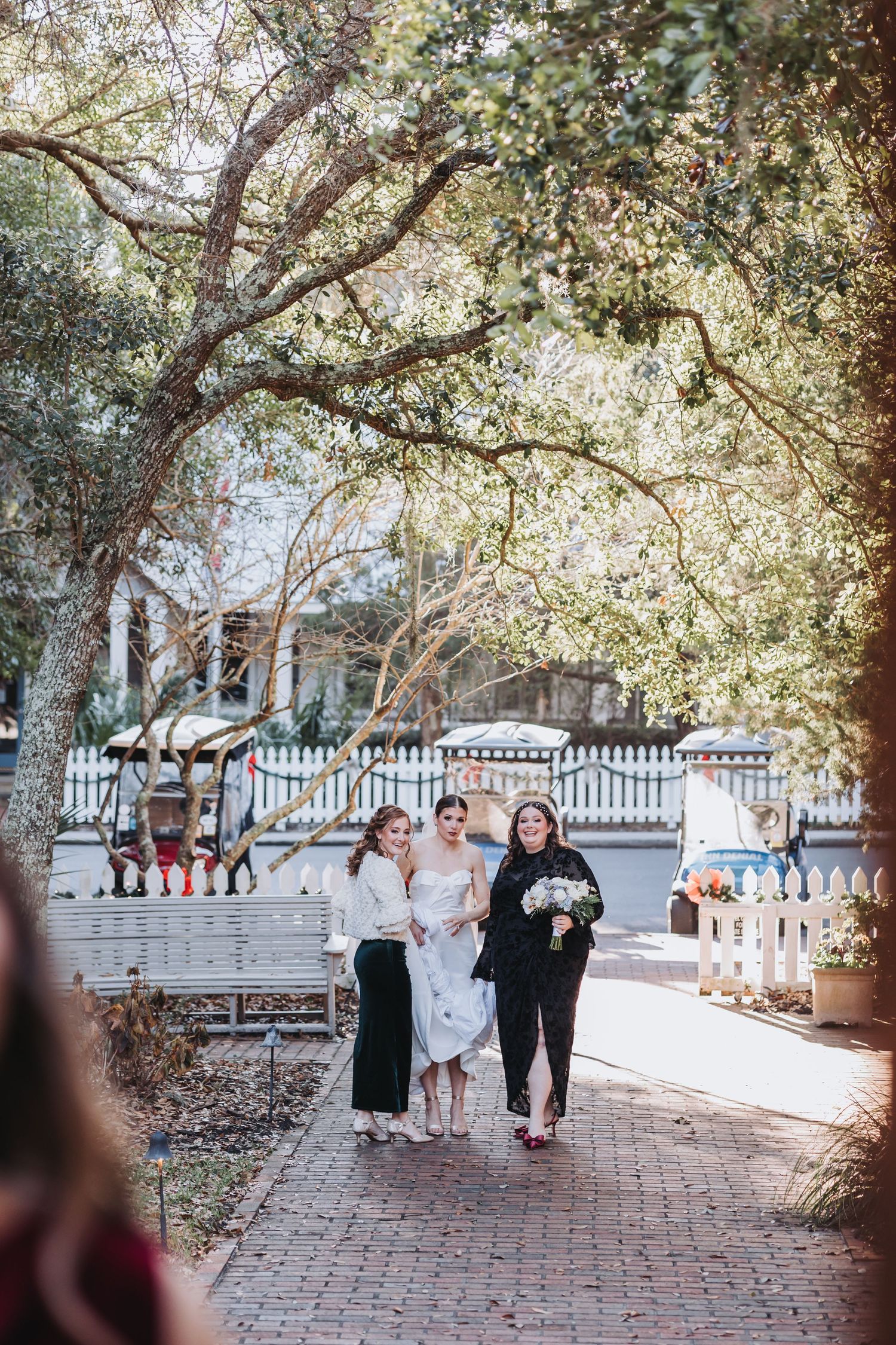 Bride in white gown walks with two women along brick pathway under oak trees toward outdoor wedding venue.
