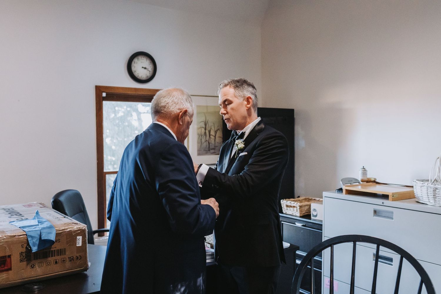 Two men in formal suits shaking hands in an office before a wedding ceremony.