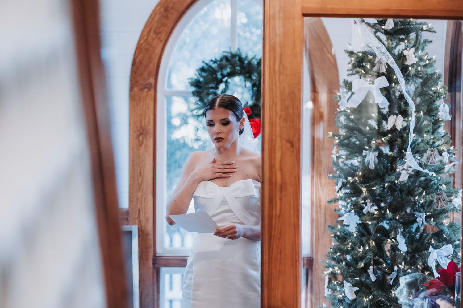 Elegant bride in strapless white gown stands in arched doorway framed by Christmas tree decorations.