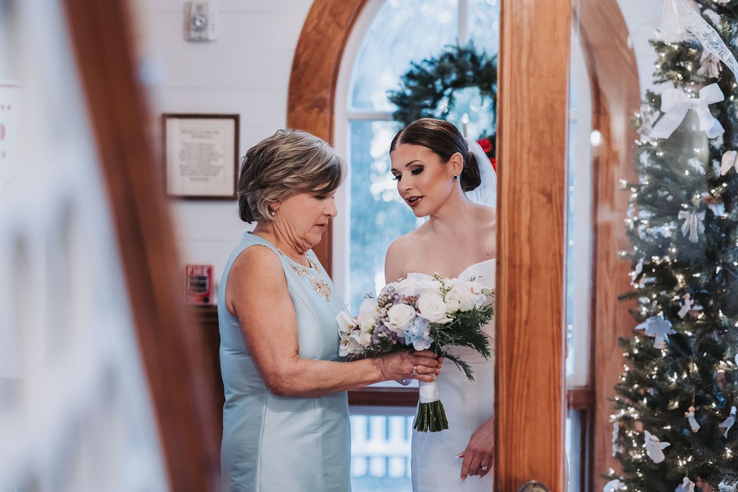 Bride in strapless white gown holds bouquet with older woman in blue dress inside decorated chapel.