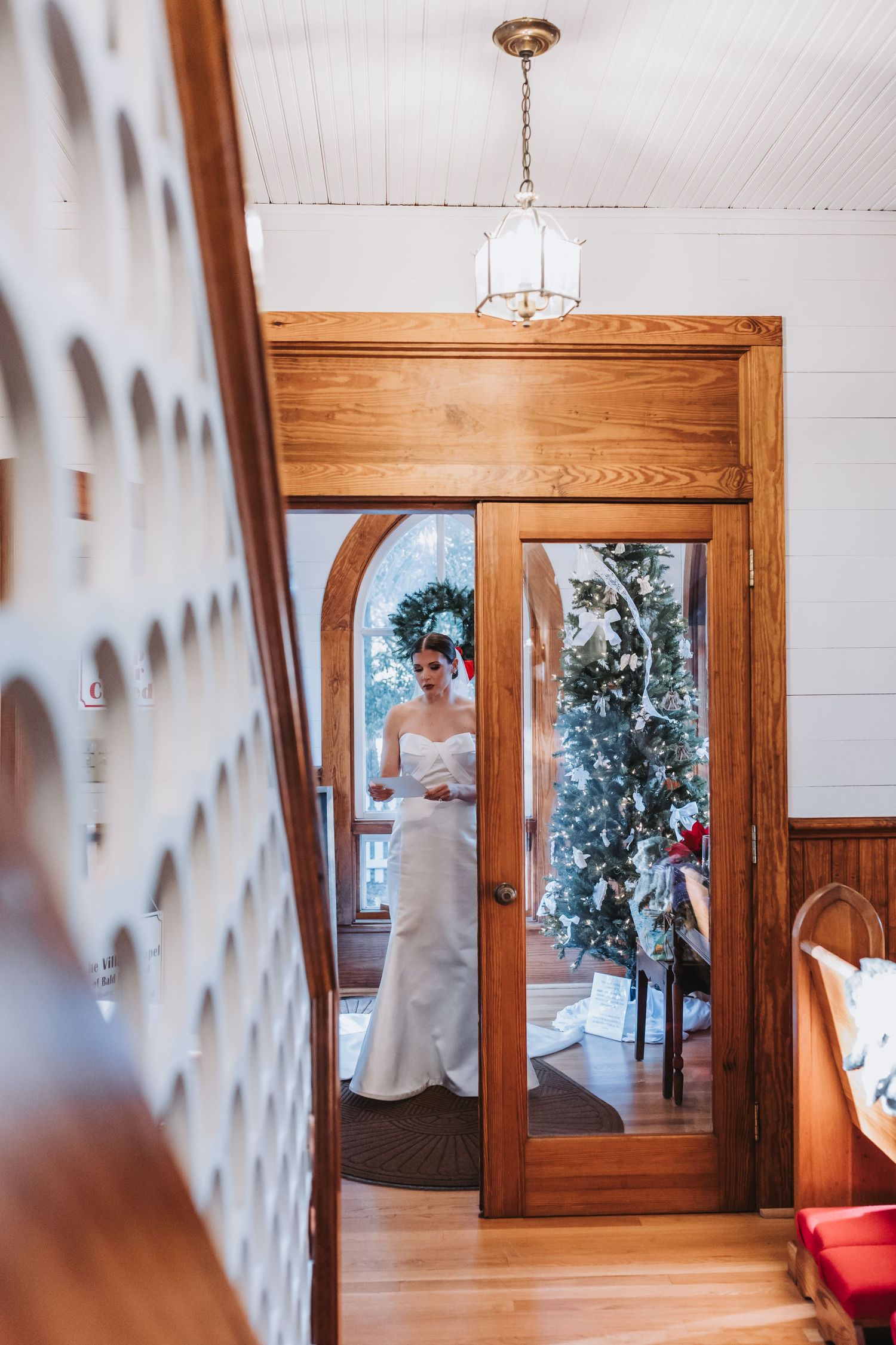 Bride in a white strapless gown standing in a doorway with a decorated Christmas tree behind her.