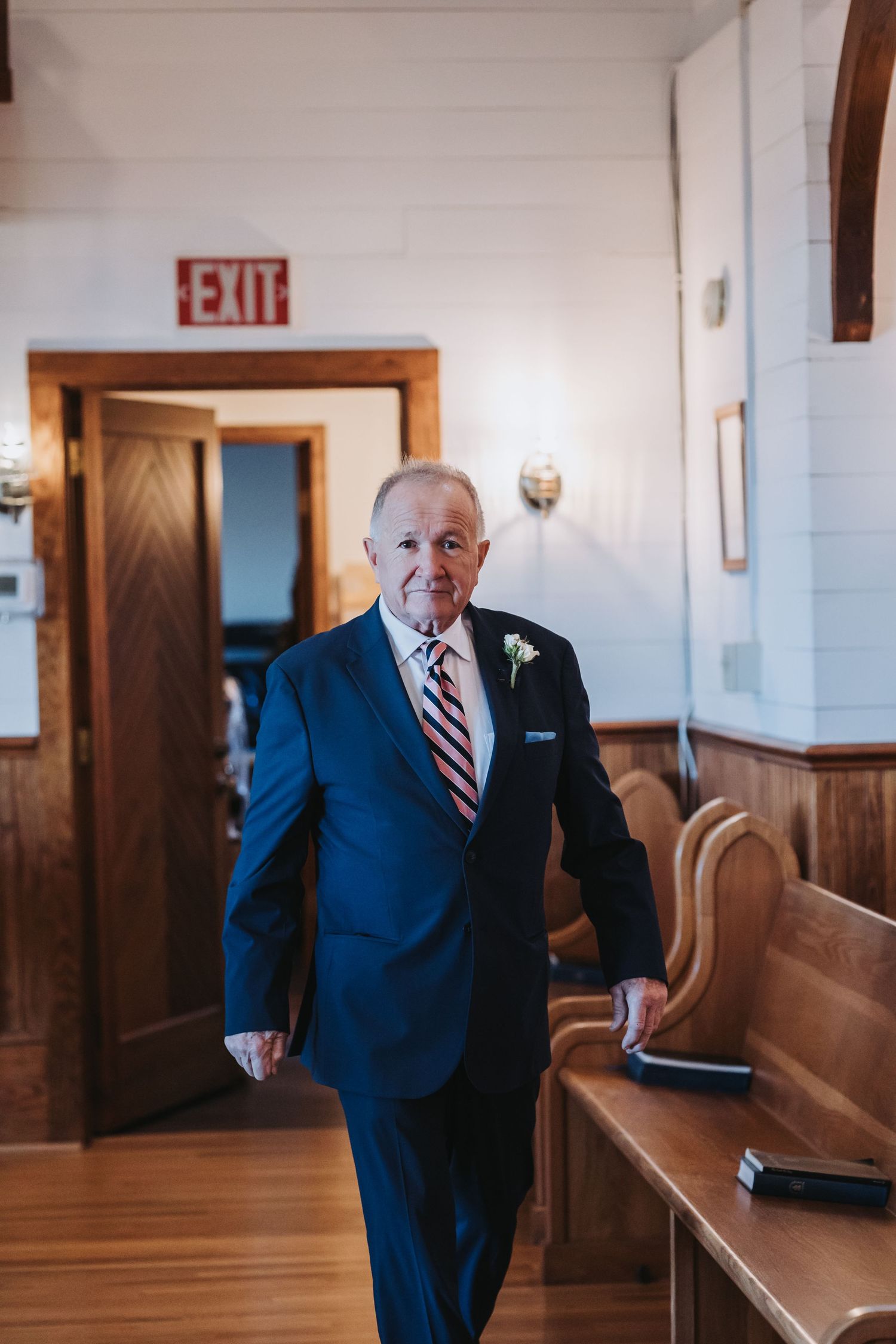 Elderly man in navy suit with boutonniere walking through a church aisle at a wedding.