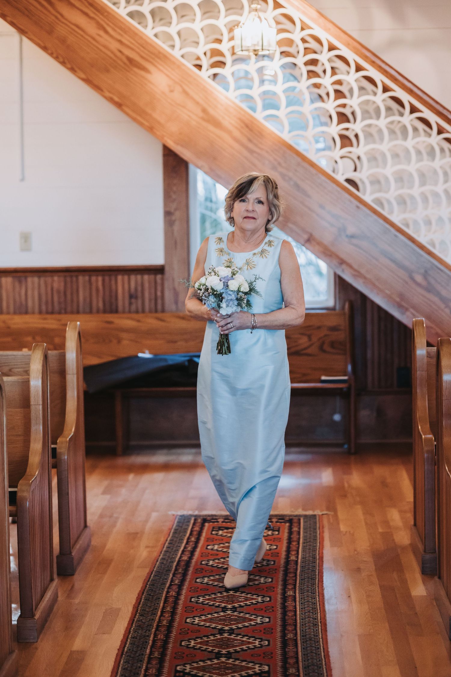 Woman in light blue gown walks down church aisle carrying white floral bouquet in charming wooden chapel.
