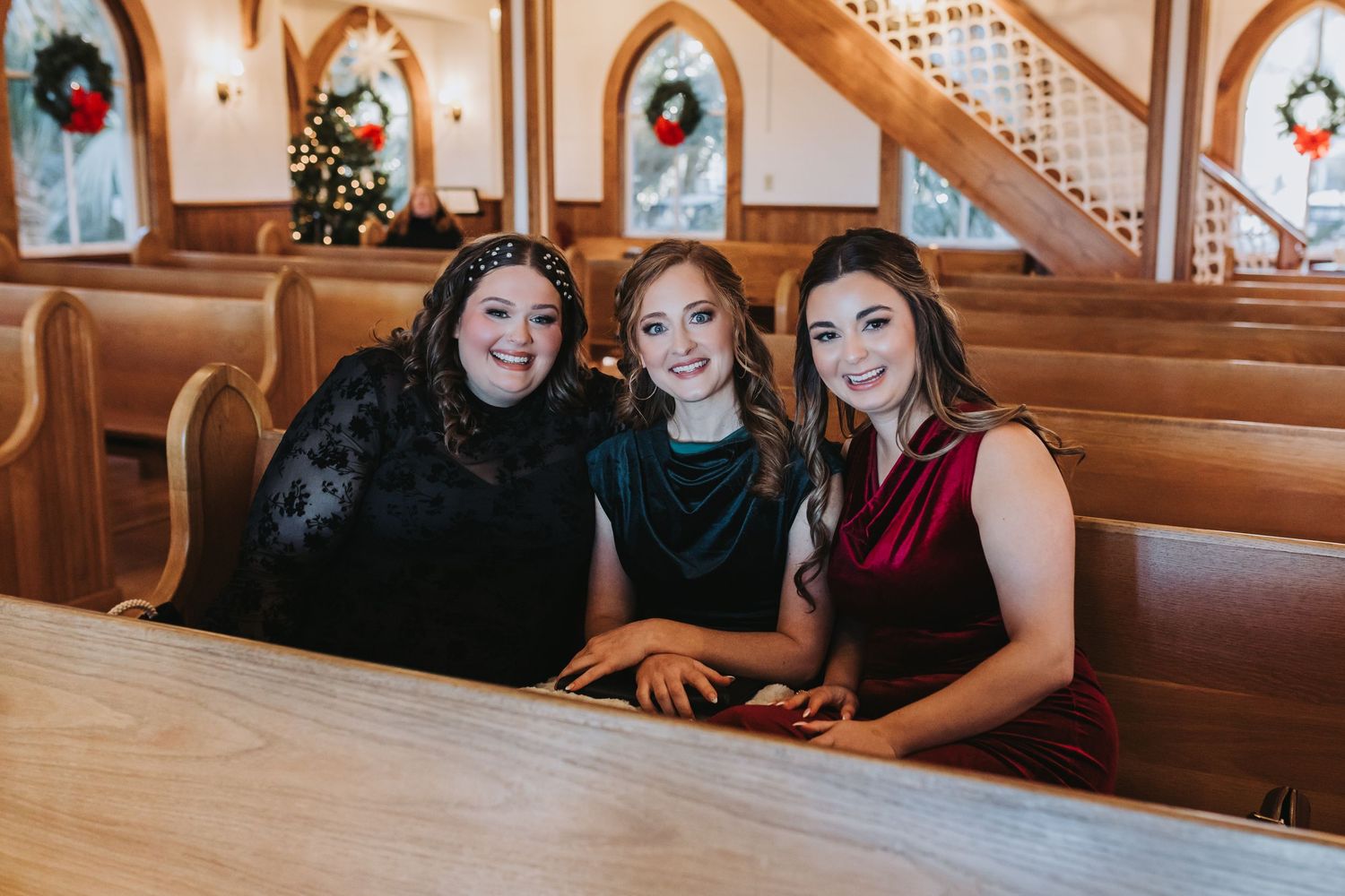 Three women in elegant dark dresses smiling together in wooden church pews with Christmas decorations.