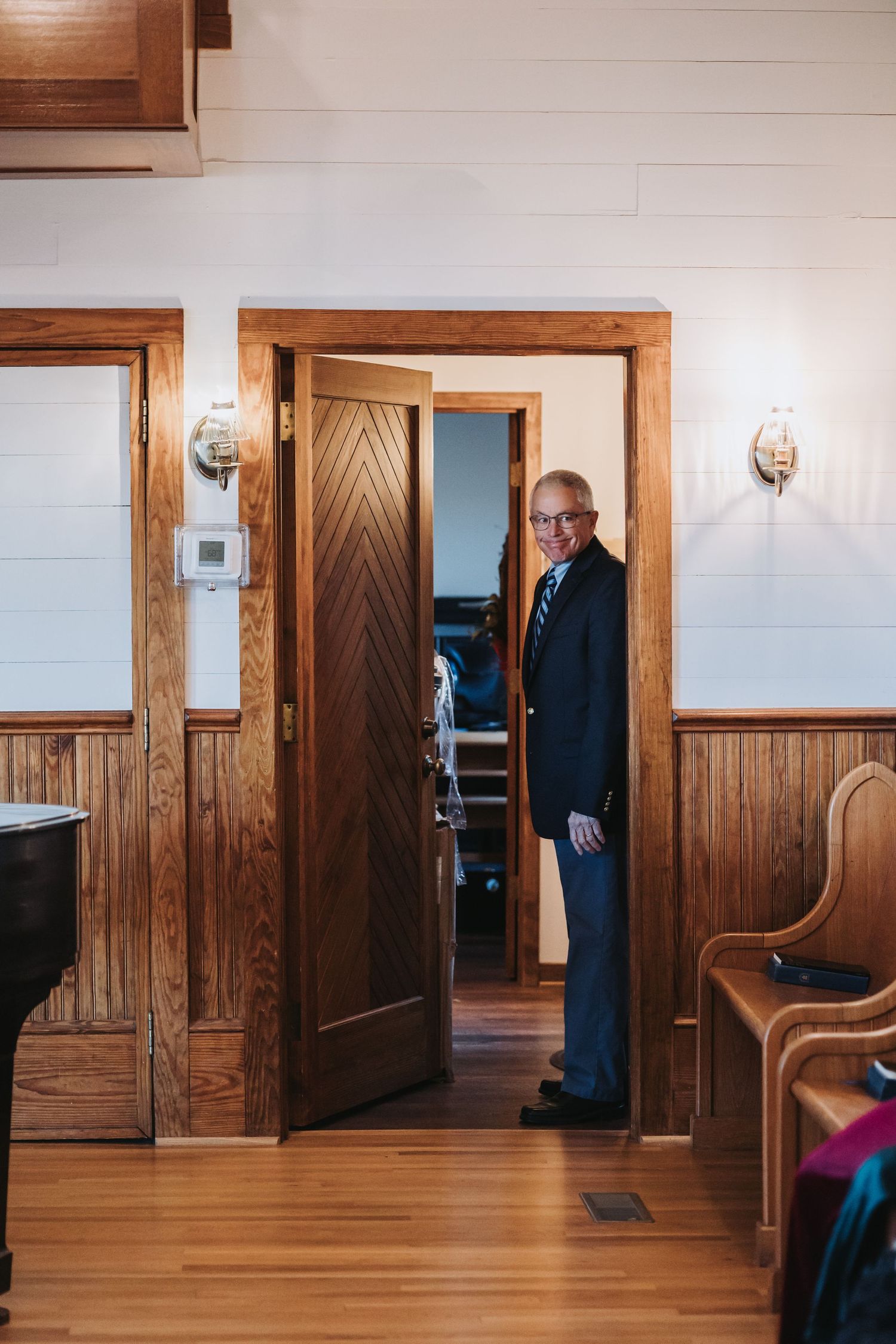 Gentleman in a navy suit smiling while standing in a wooden doorway of a rustic chapel.