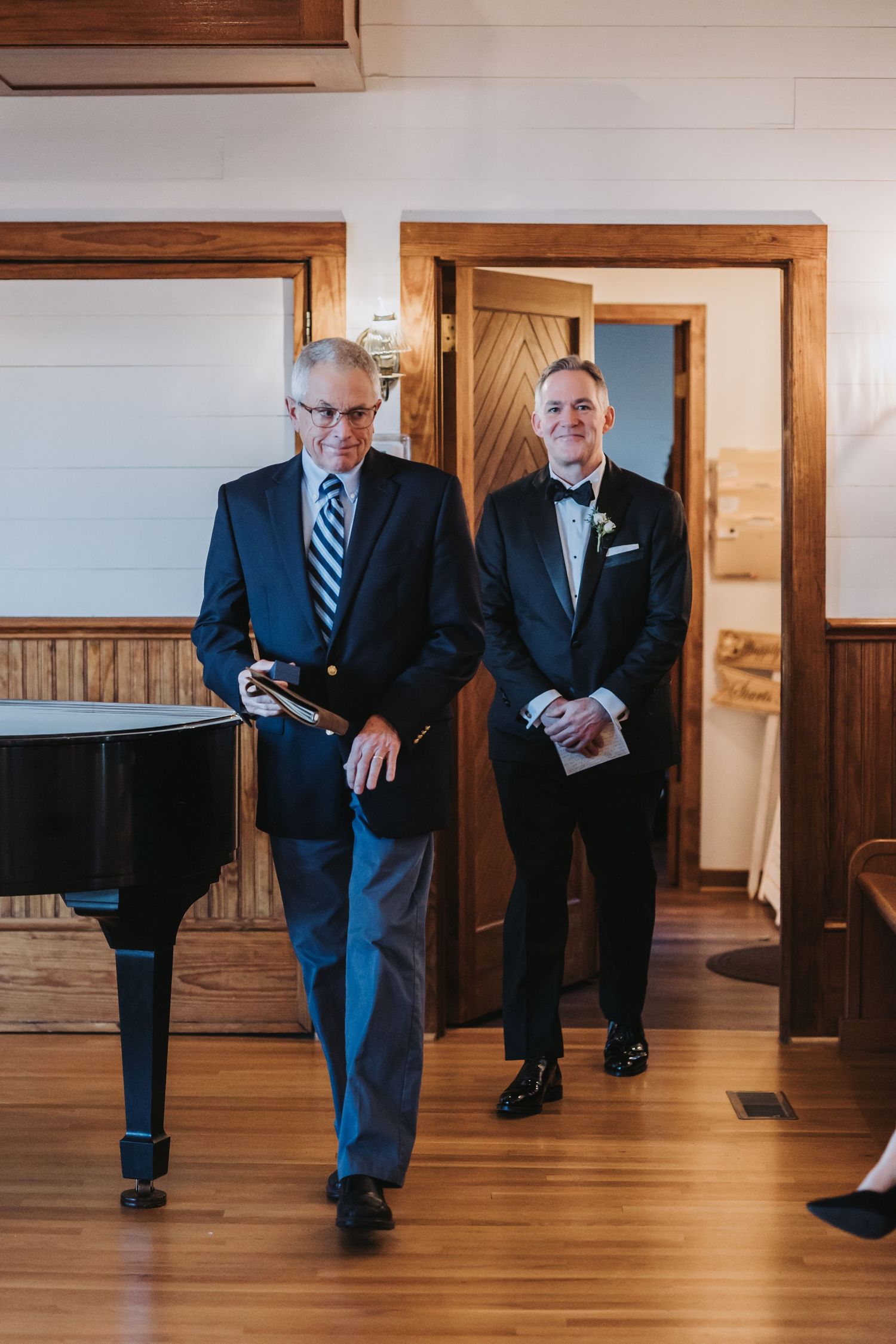 Groom in black tuxedo smiles as he walks down aisle with older man in navy suit at wedding ceremony.