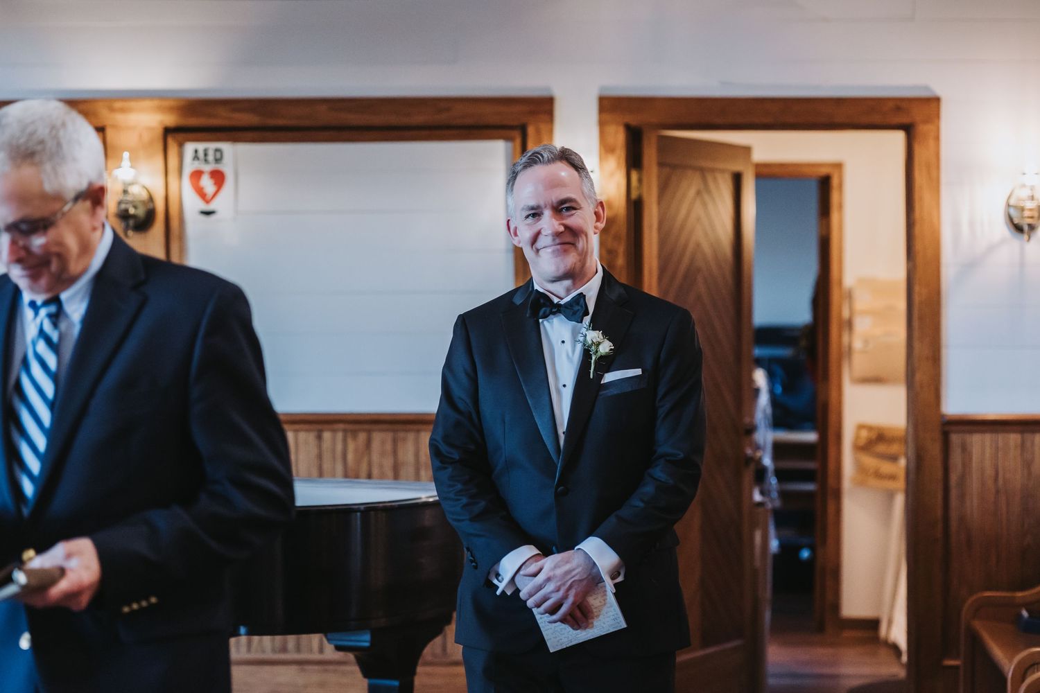 Smiling groom in black tuxedo with boutonniere awaits bride during indoor wedding ceremony.