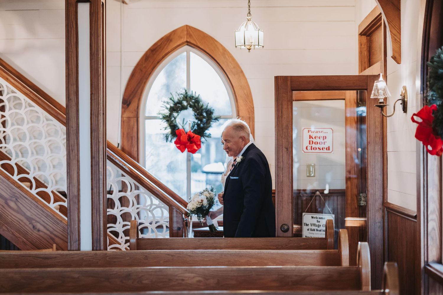 Elderly man in suit carries white bouquet through wooden church interior decorated with Christmas wreaths.