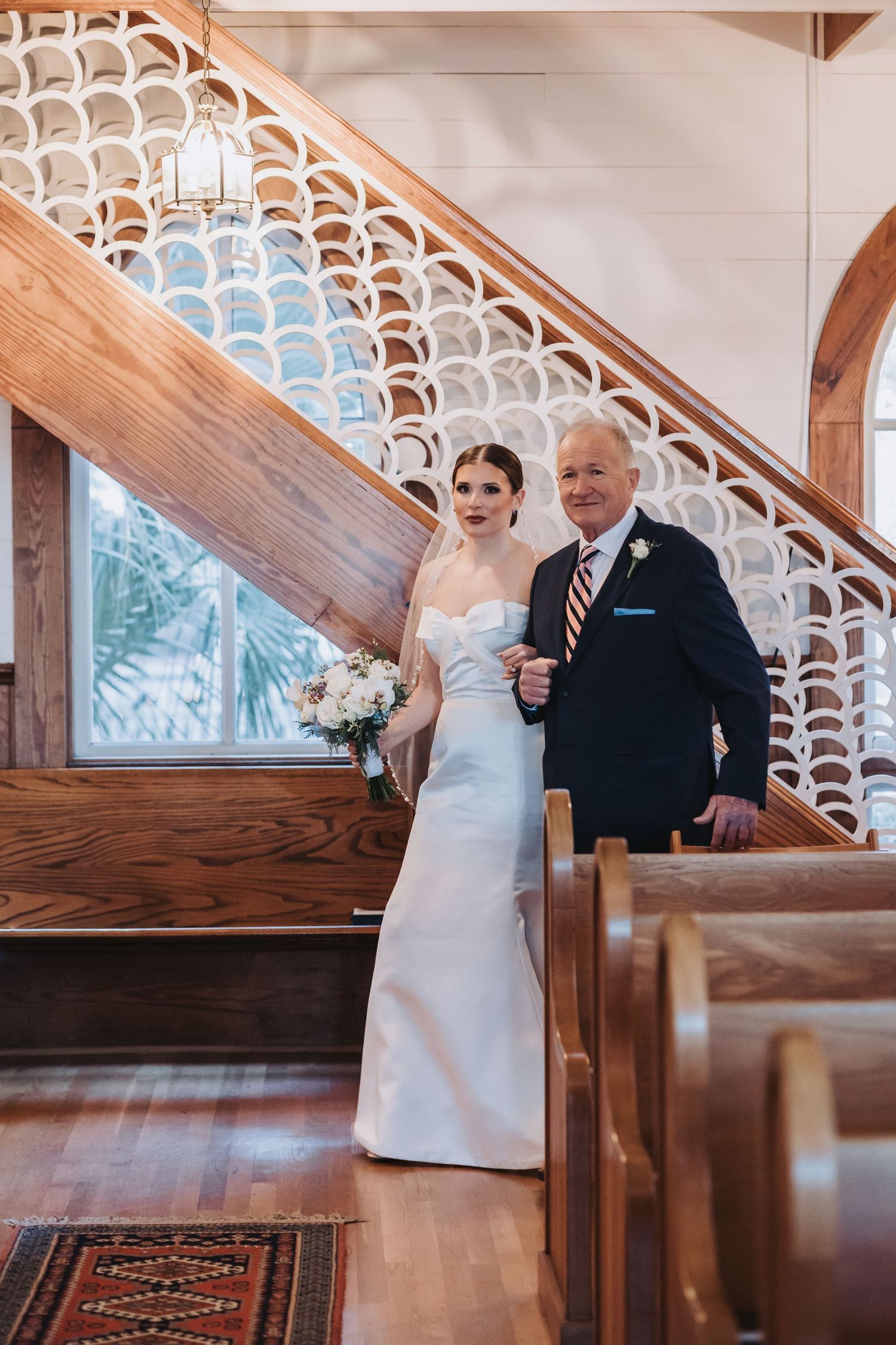 A bride in a white gown walks down the aisle with an older man in a navy suit inside a wooden church.