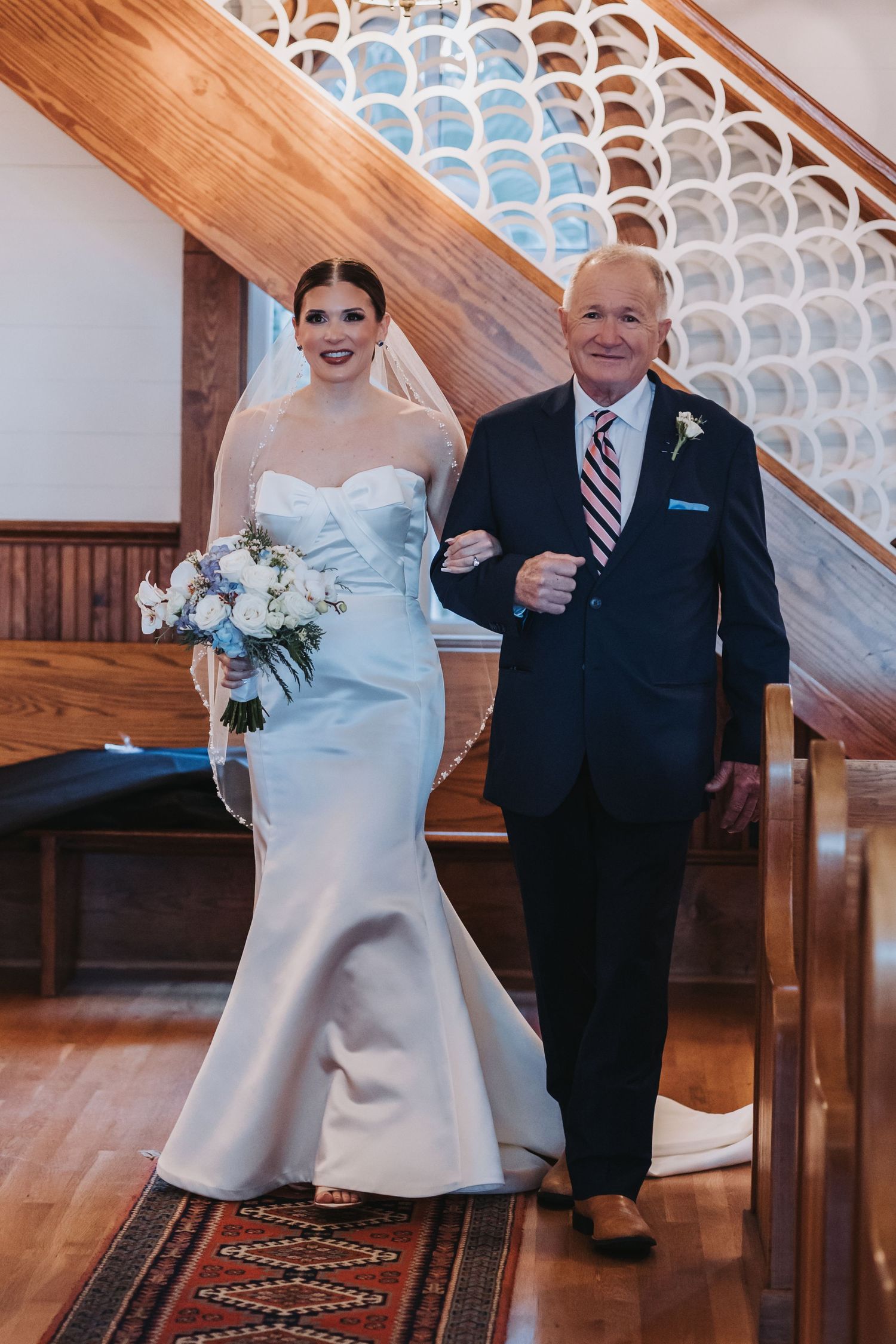 Bride in strapless white gown walks down aisle with older gentleman escort in navy suit.