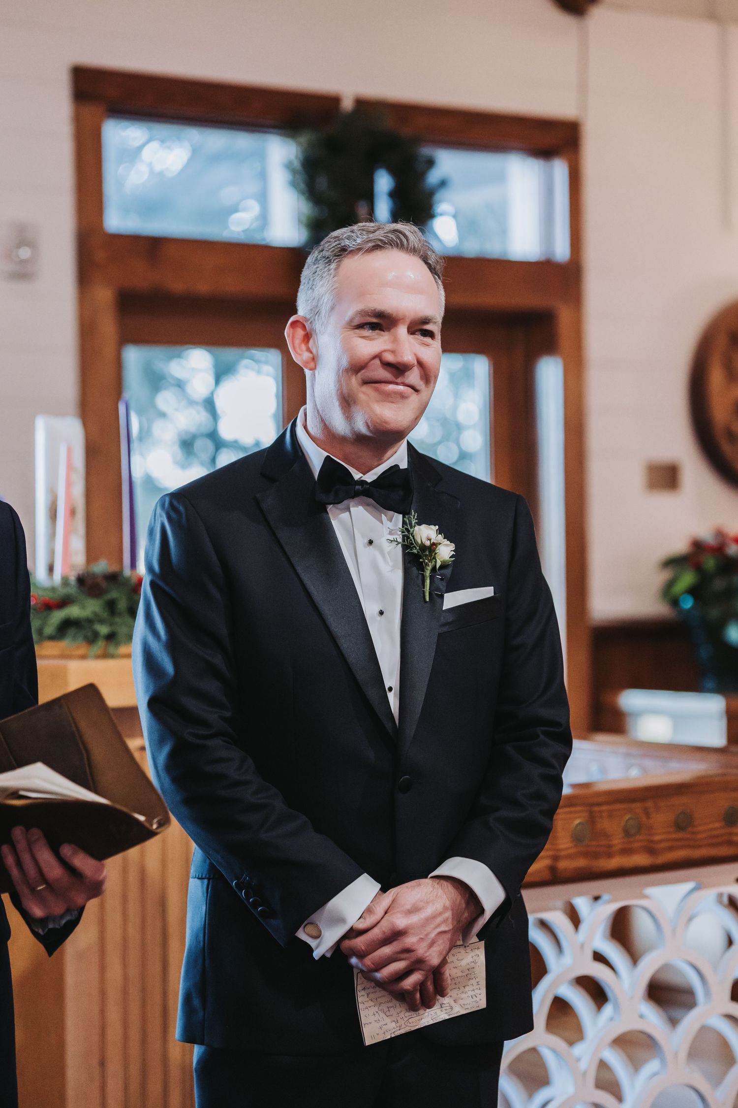 Emotional groom in black tuxedo smiles warmly while waiting at the altar during wedding ceremony.