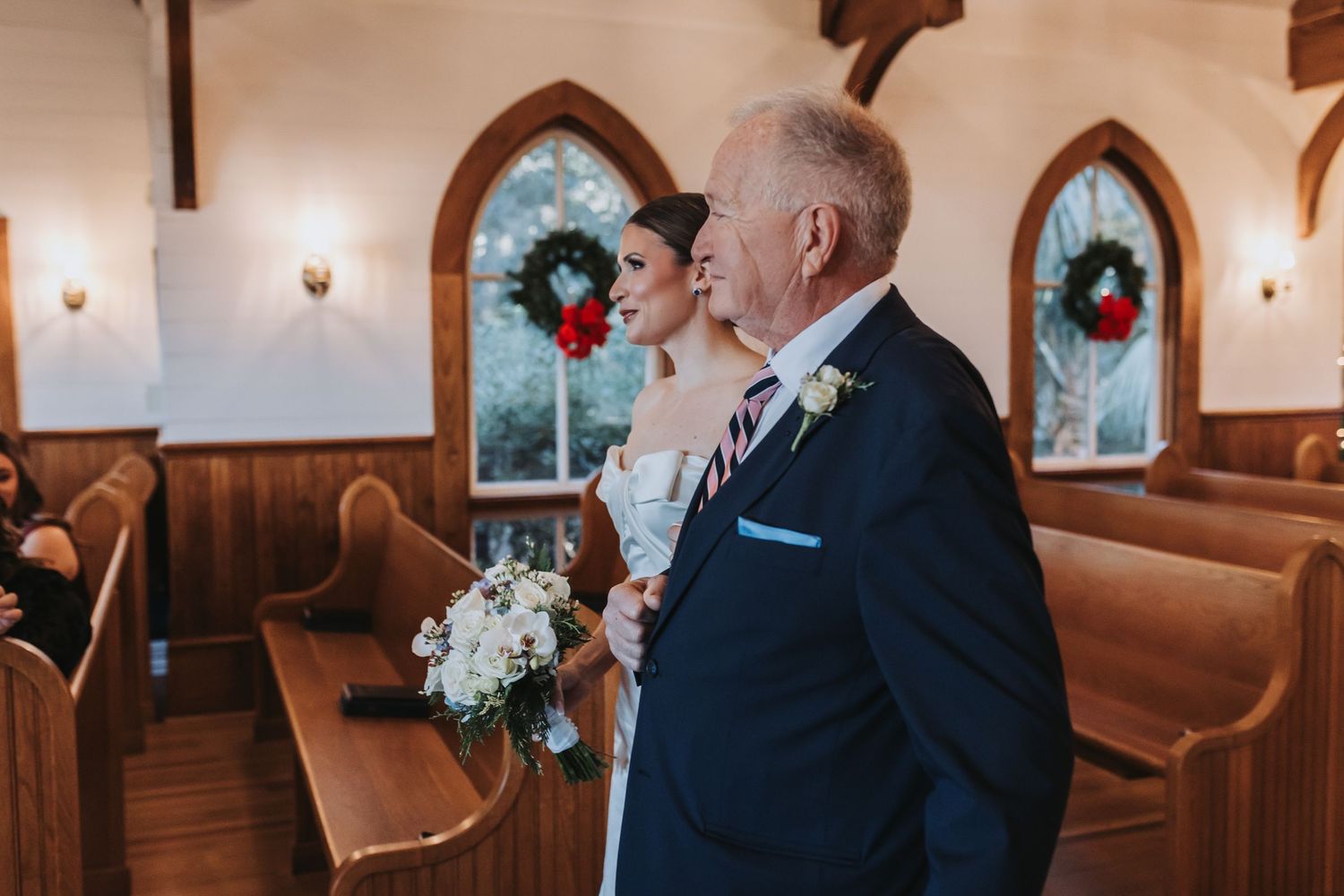 A bride holding a white bouquet walks down the church aisle with an older gentleman in a navy suit.