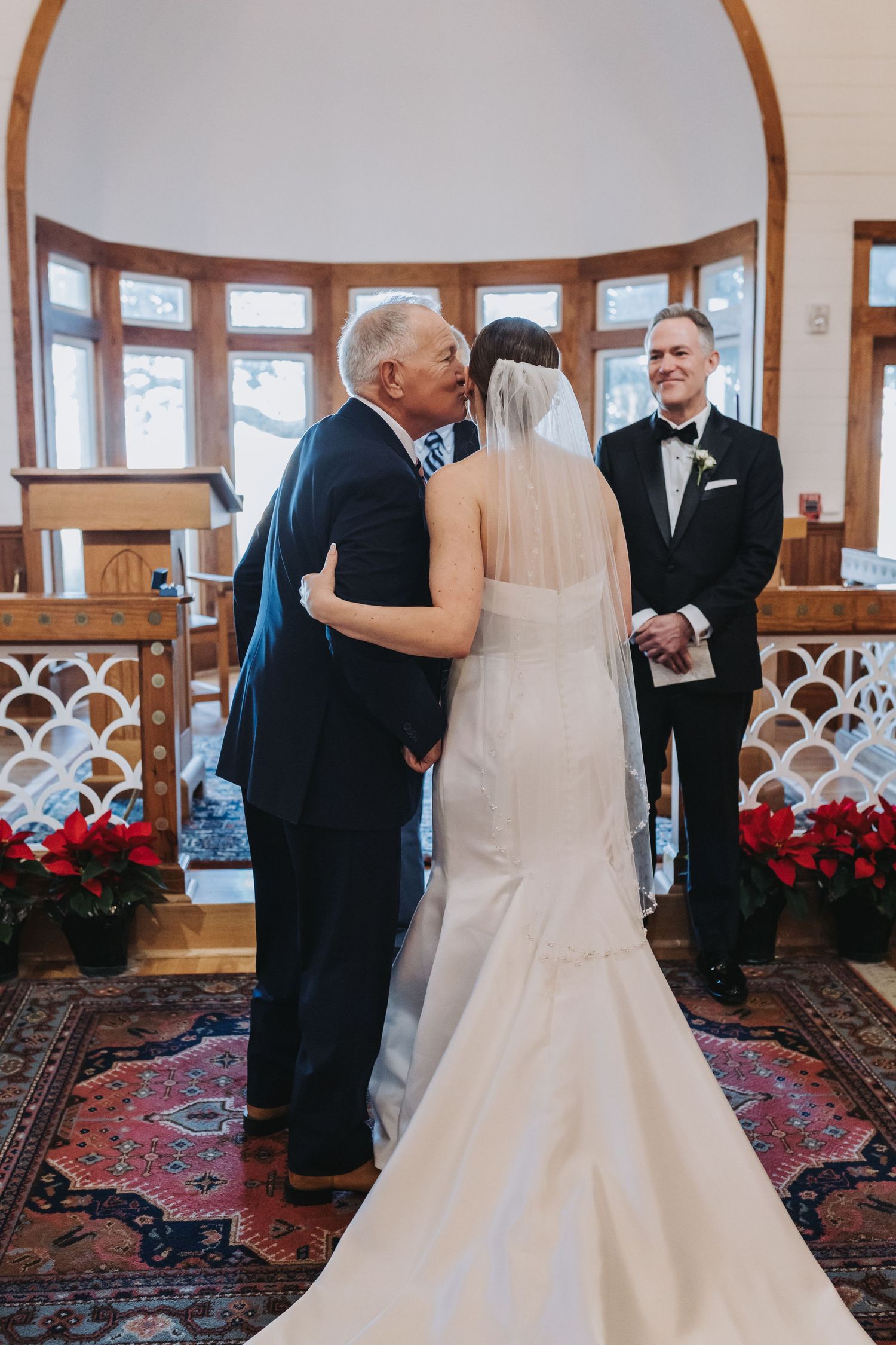 Father kisses bride at wedding altar while groom smiles in chapel with red floral arrangements.