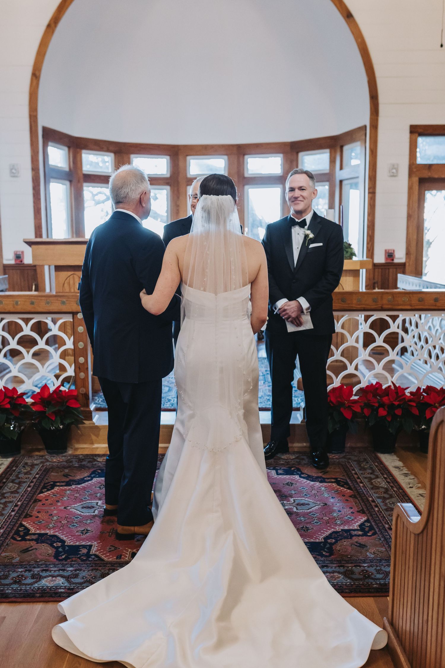 Bride and groom exchange vows at elegant chapel altar adorned with red poinsettias and arched windows.