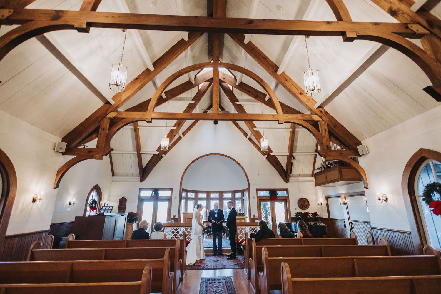 Wide view of intimate wedding ceremony inside rustic chapel with exposed wooden beams and Christmas wreaths.