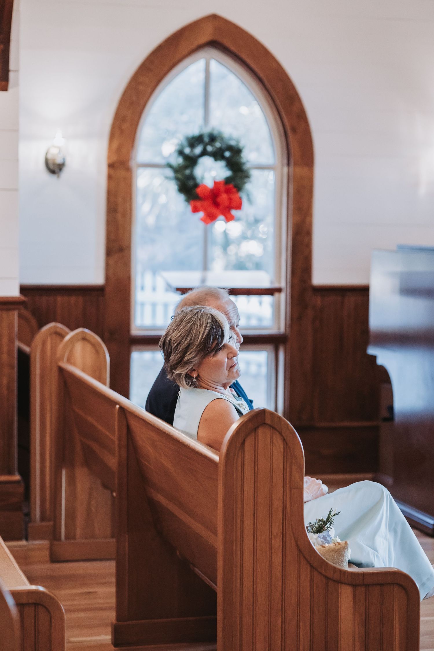 Bride in white gown sits in wooden church pew beneath Gothic arched window adorned with Christmas wreath.