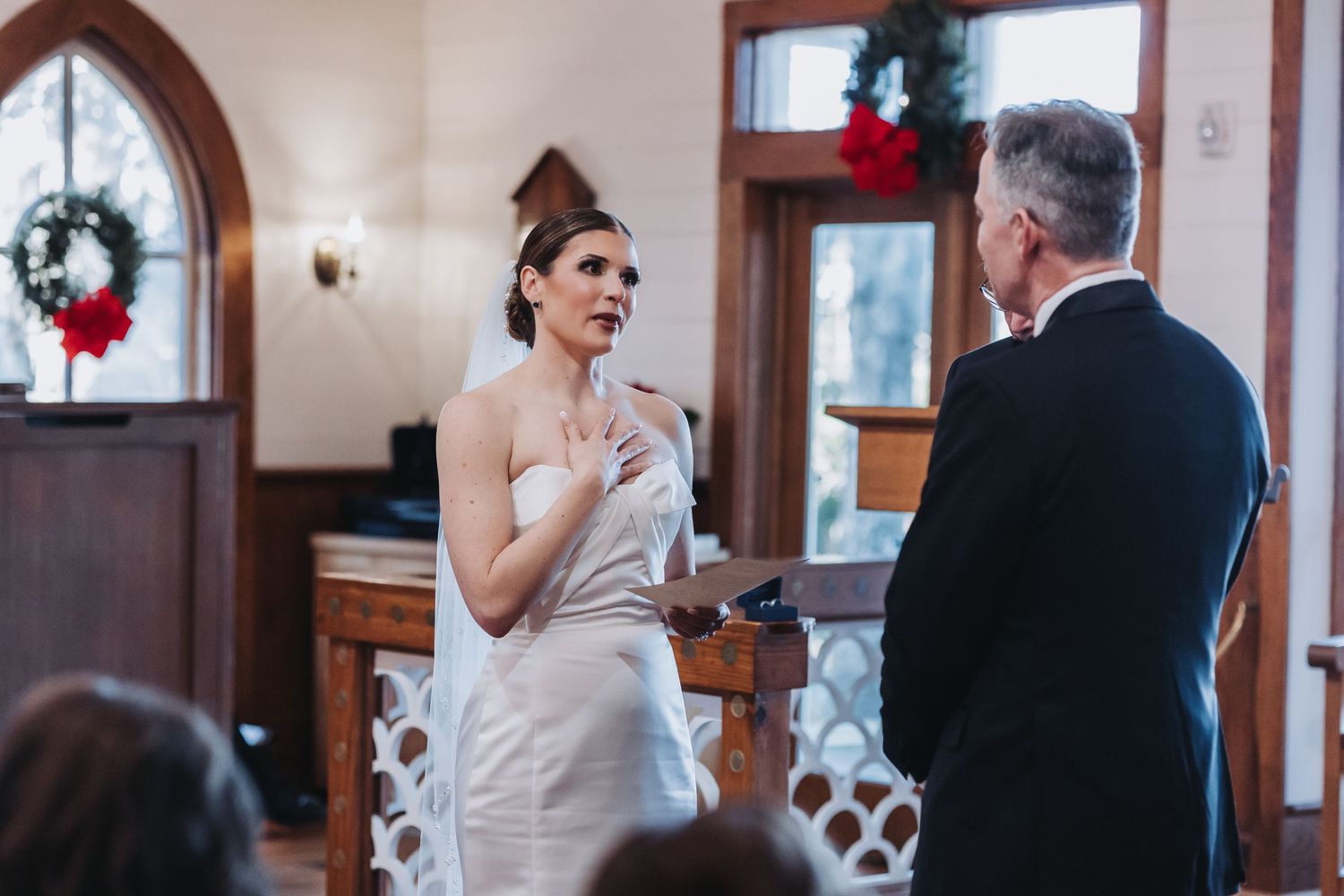 Bride in strapless white gown recites heartfelt vows to groom during intimate chapel wedding ceremony.