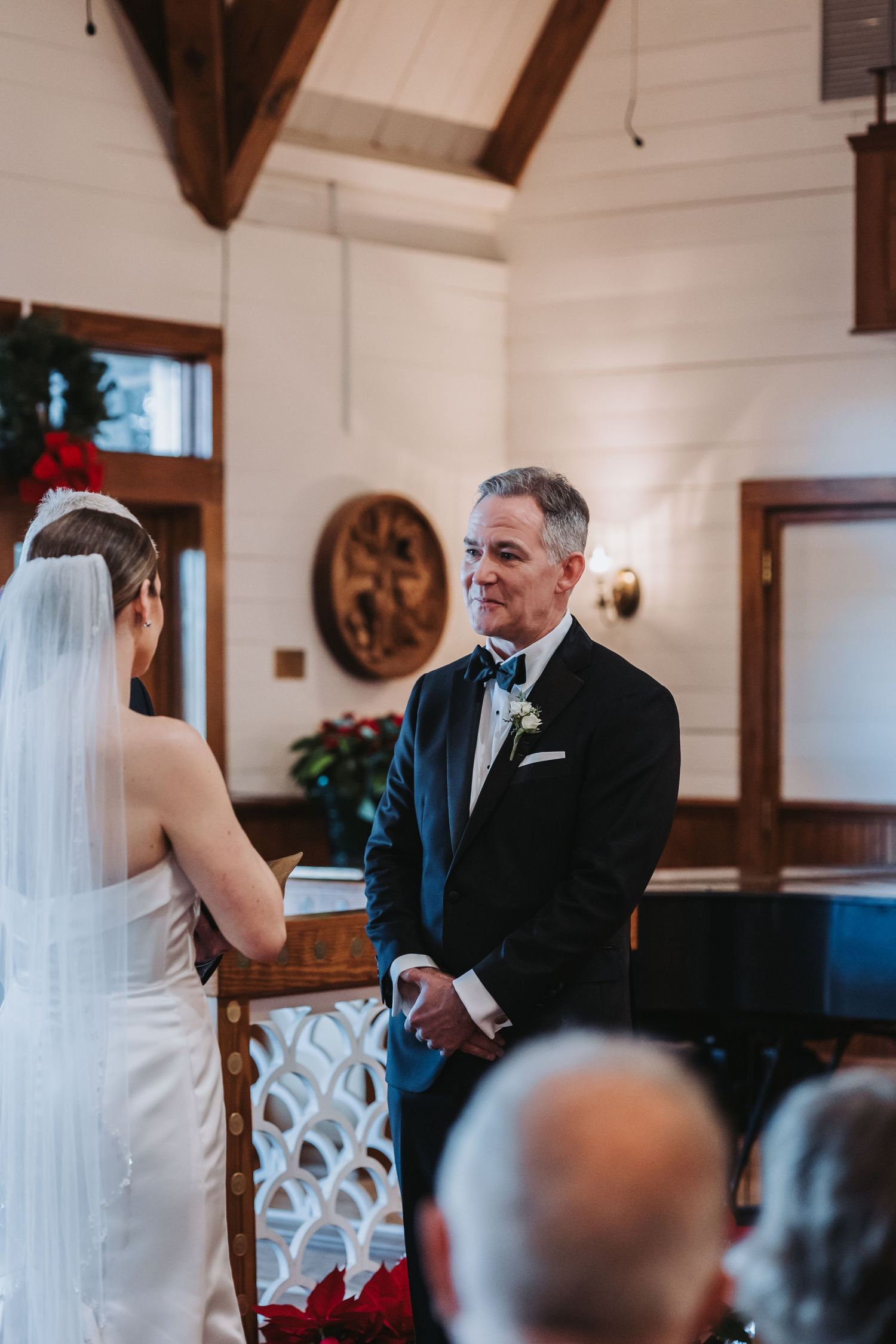 Groom in black tuxedo smiles warmly at veiled bride during intimate wedding ceremony in white chapel.