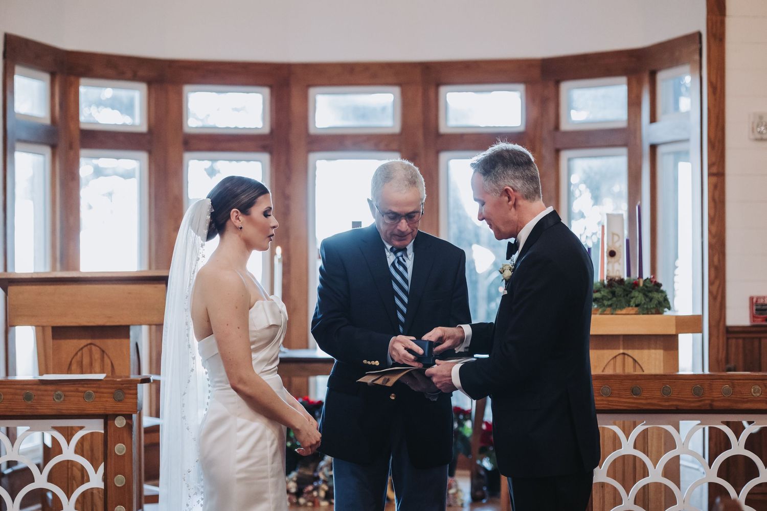 Bride and groom exchange rings with officiant during elegant chapel wedding ceremony.