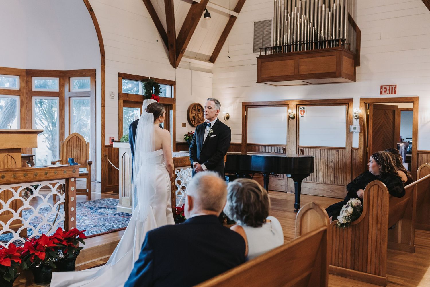 Wedding ceremony in charming chapel with pipe organ, guests seated in wooden pews watching couple at altar.
