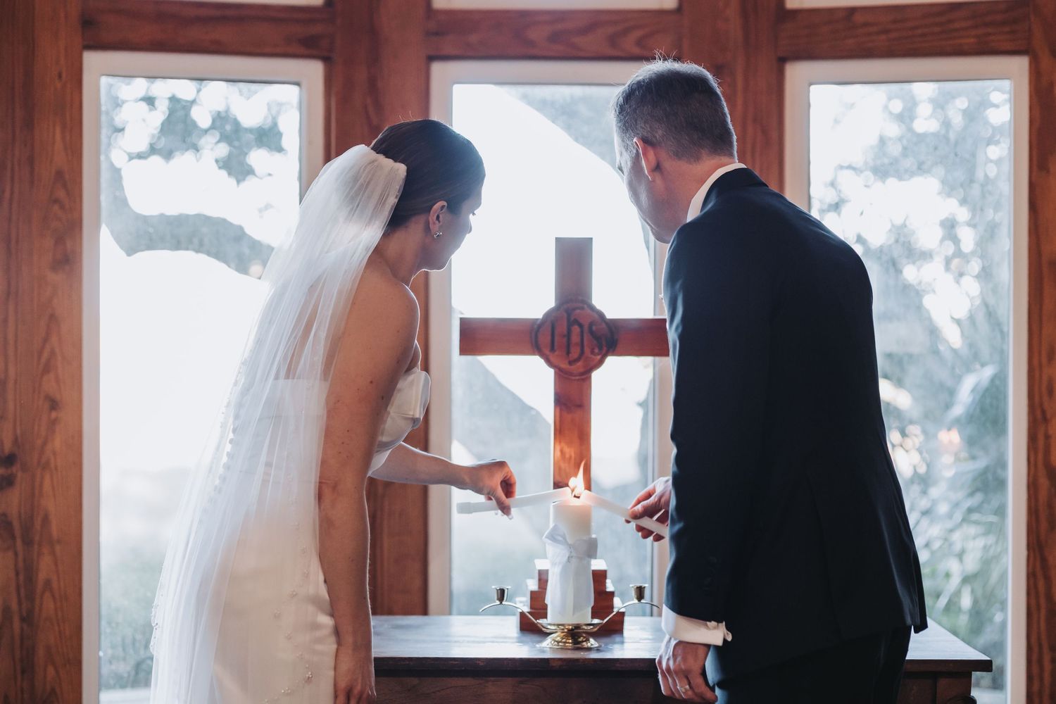 Bride and groom lighting unity candle before wooden cross in chapel window during wedding ceremony.