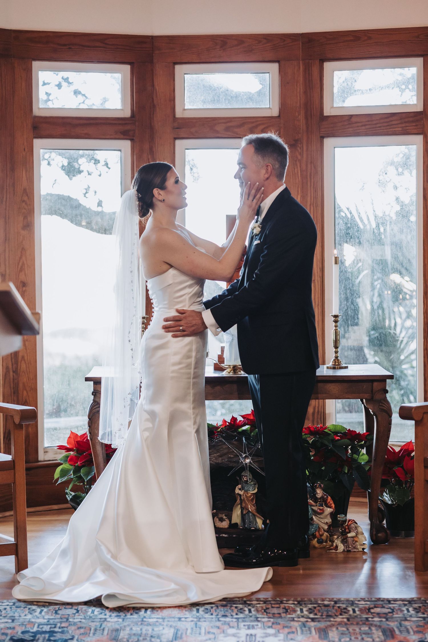 A bride in a white gown tenderly holds the face of her groom near a festive holiday altar.